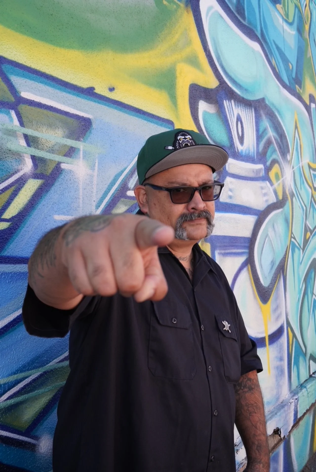 A man with sunglasses and a cap pointing toward the camera, standing in front of a colorful graffiti wall.