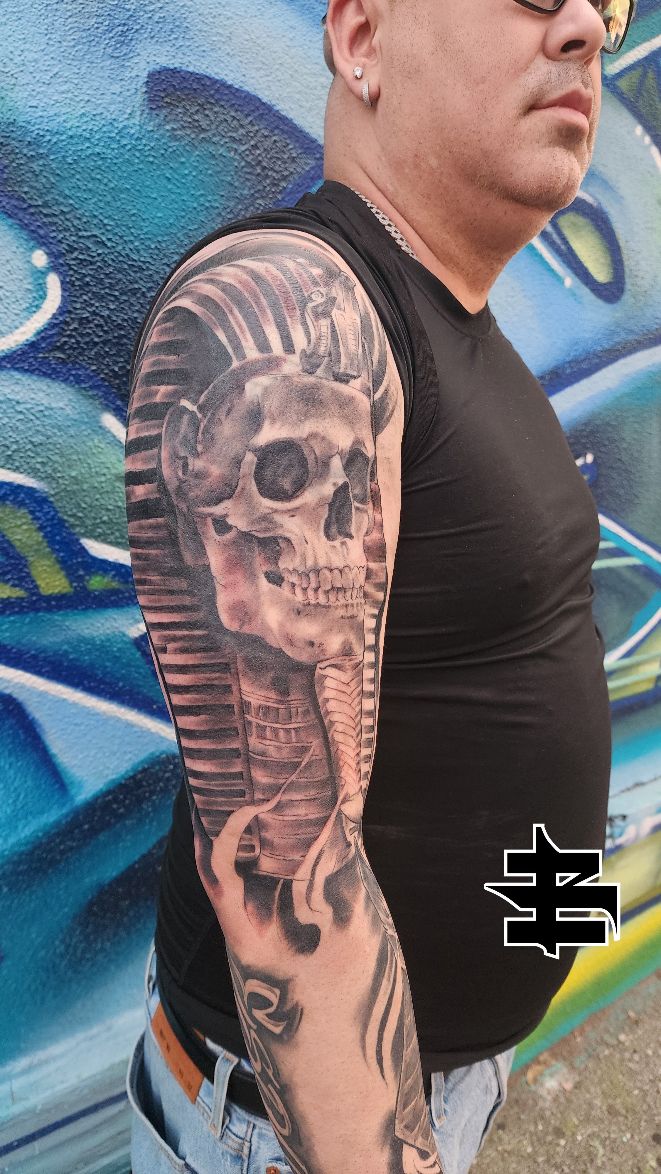 A man with a large tattoo of a skull wearing a pharaoh headdress on his upper arm, standing against a graffiti wall.