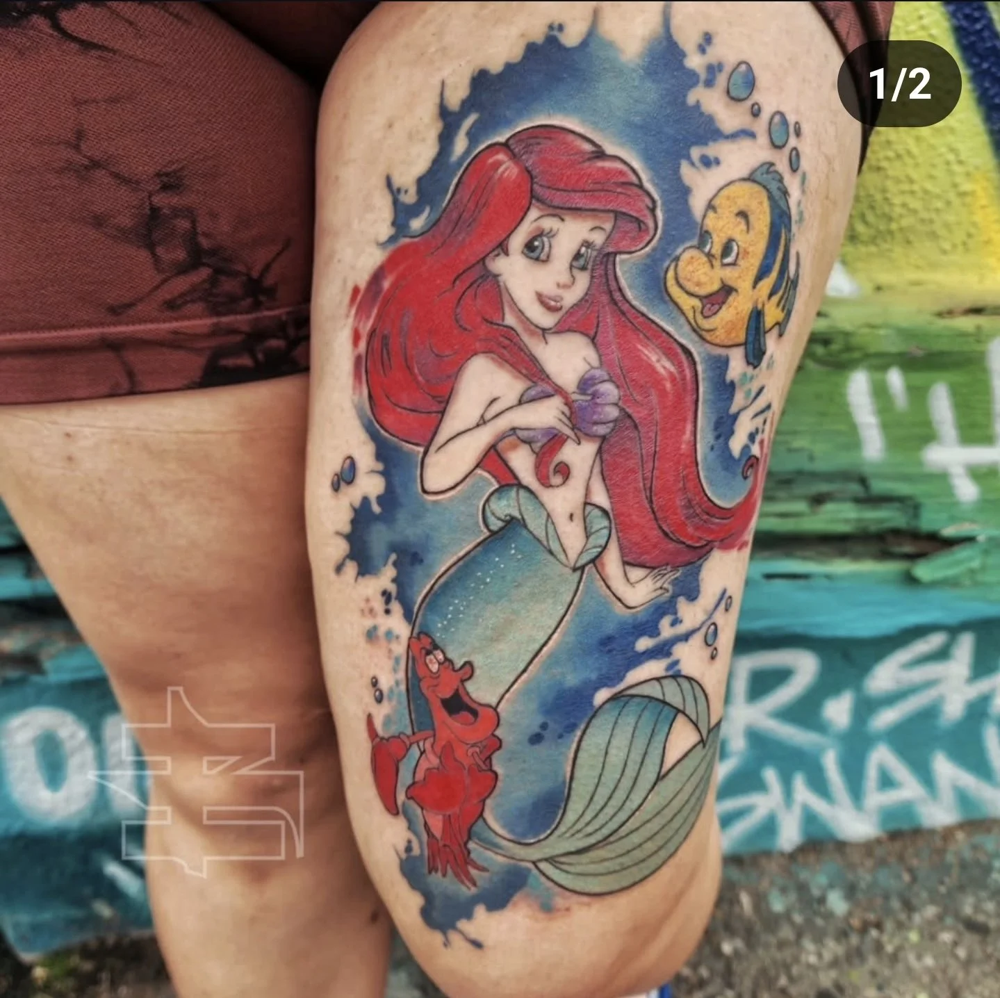 Colorful tattoo on person's arm featuring Ariel, the red-haired mermaid from Disney, with Flounder, the yellow and blue fish, and Sebastian, the red crab.