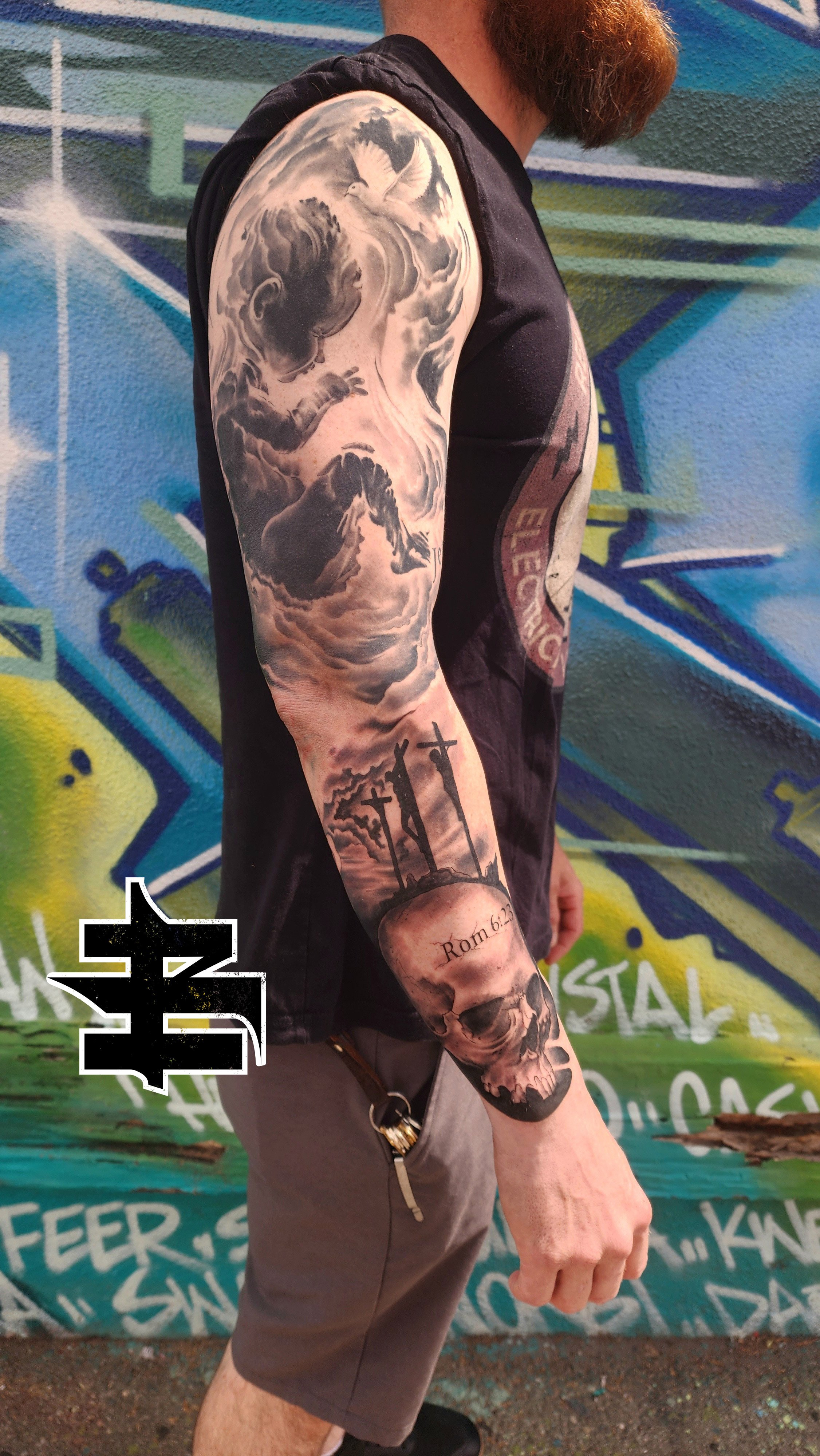Tattooed arm of a man with a large tattoo of a praying mantis, stormy clouds, a dove, and a landscape with three crosses and a skull, with the text 'Romans 6:23' on the forearm, standing against a colorful graffiti wall.
