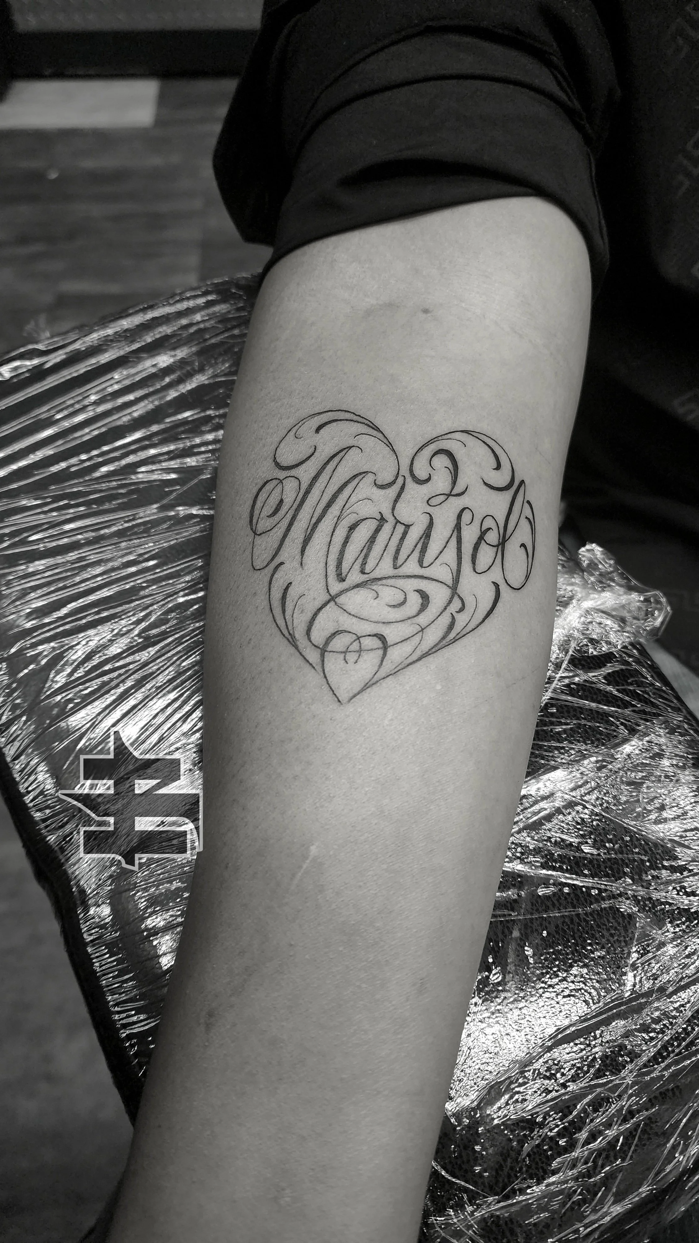 A tattoo on a person's forearm that reads 'Marisol' in elegant, cursive script, with decorative swirls. The person is sitting in a tattoo chair, covered with a metallic plastic sheet.