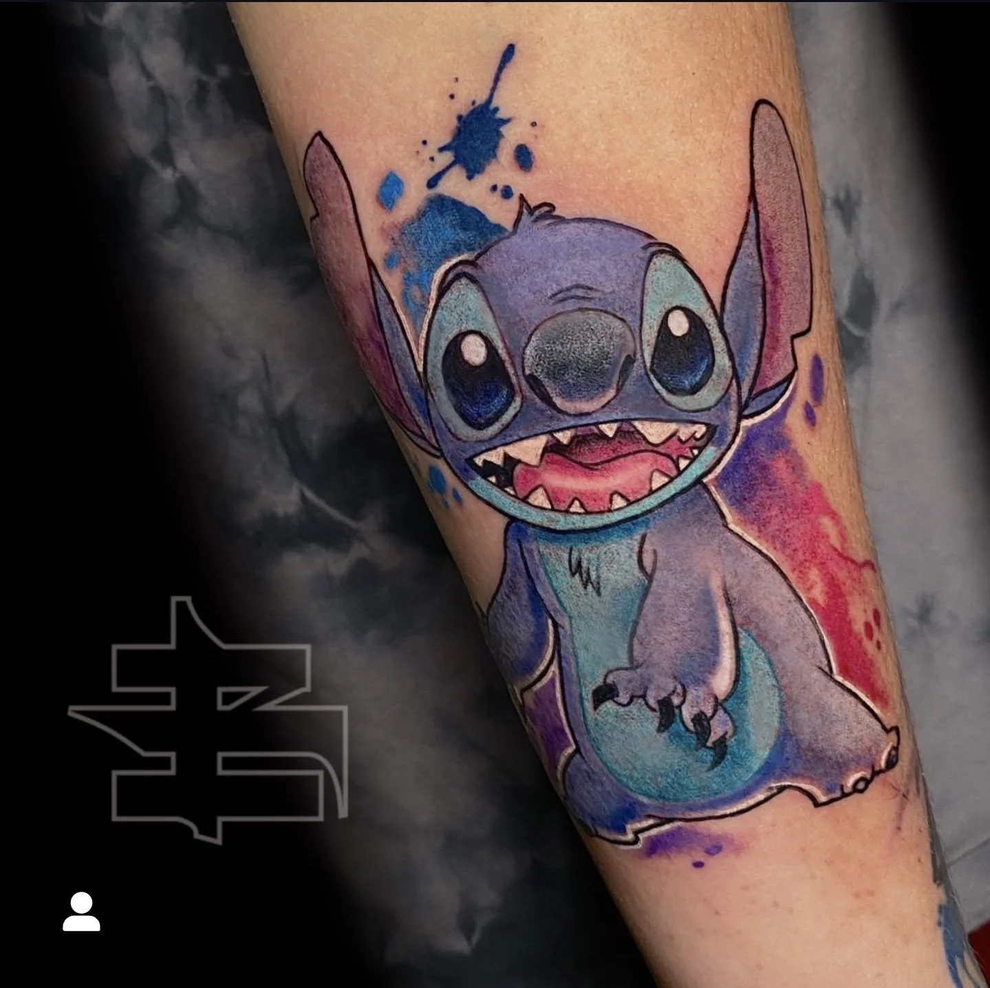Colorful tattoo of Stitch, the cartoon alien character from Disney's Lilo & Stitch, with a splash of blue ink on its forehead and vibrant accents on its ears and body.