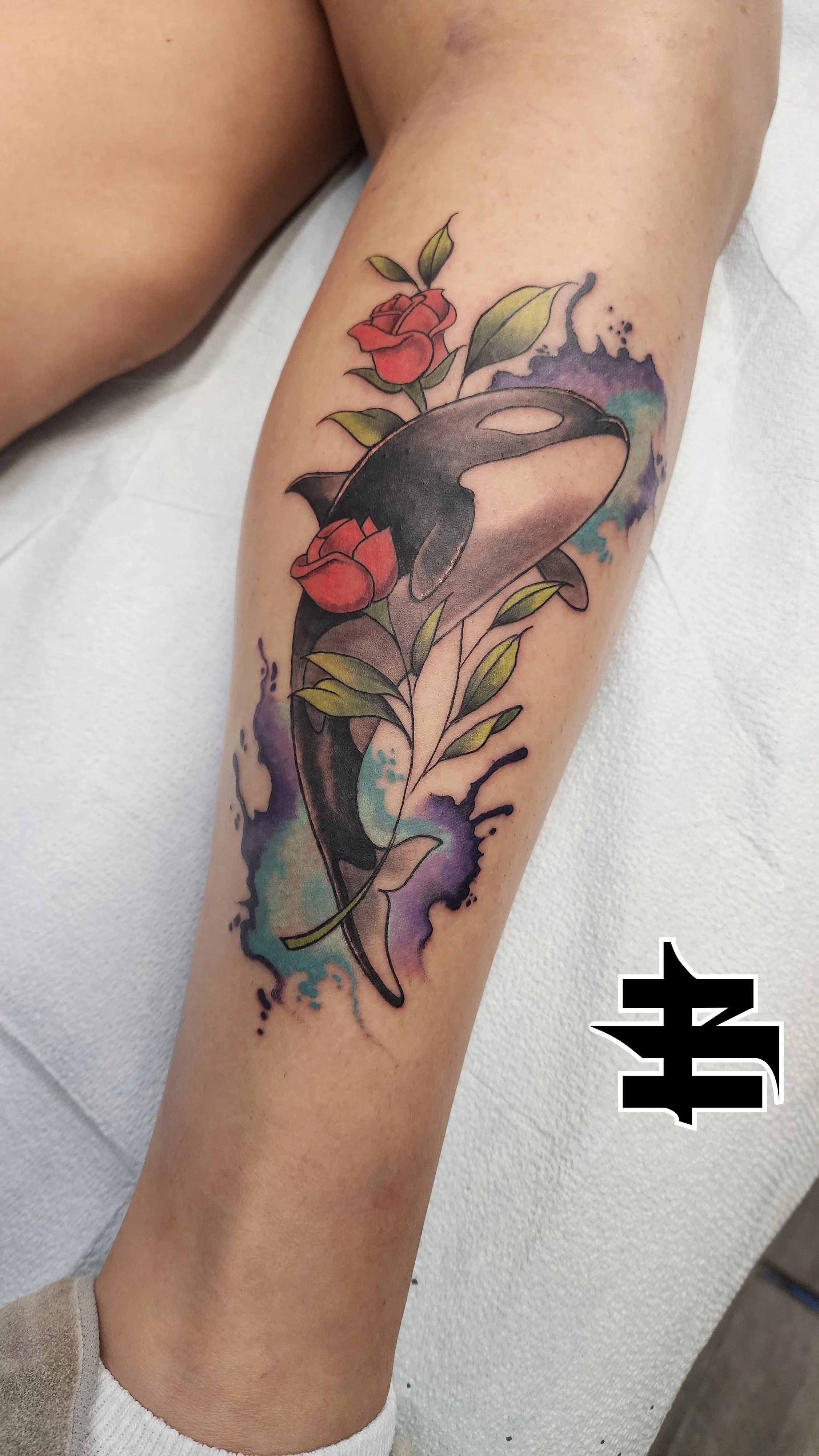Tattoo of a whale with red roses and green leaves on a person's arm, with colorful watercolor splash background.