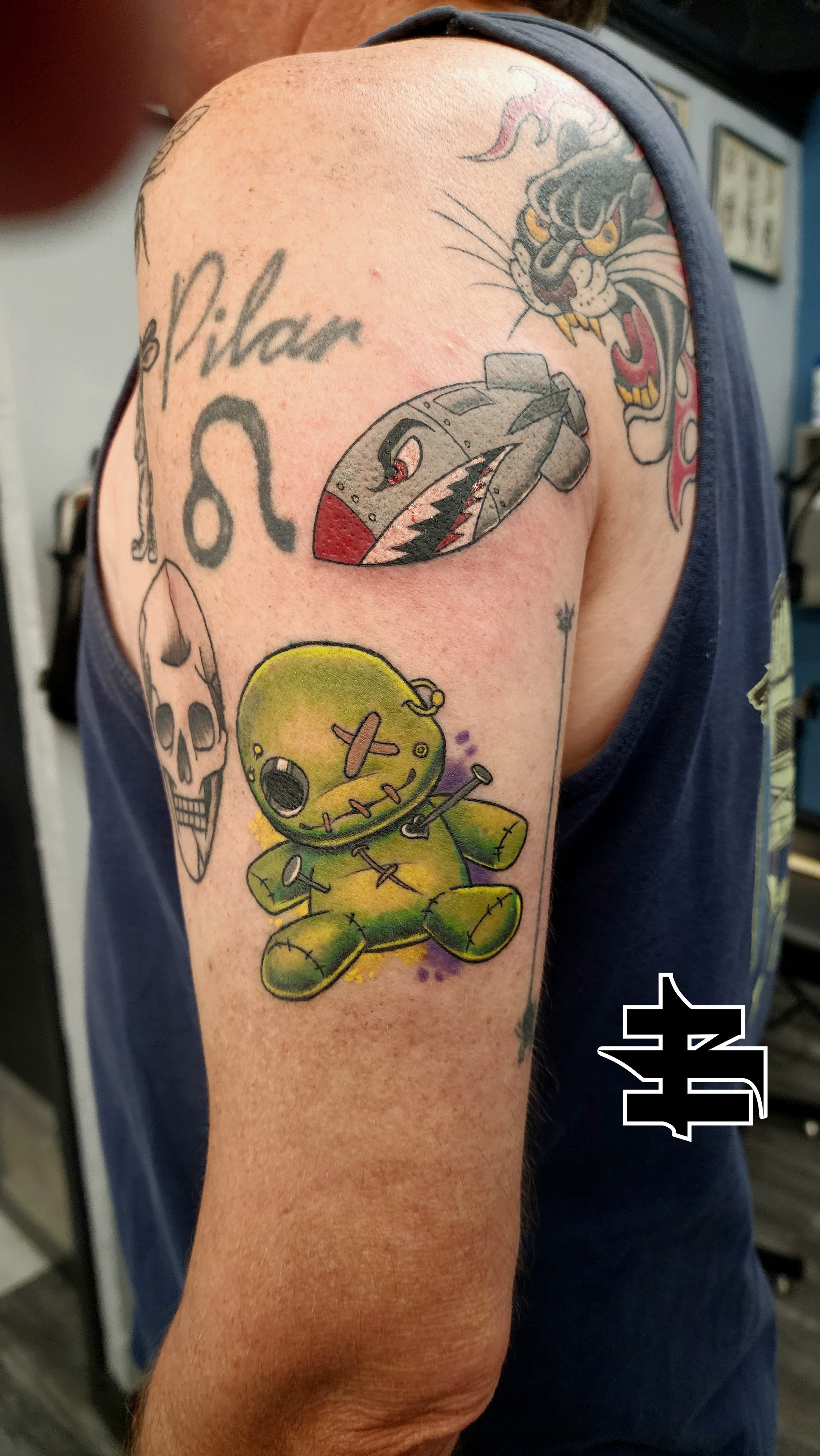 Tattoo of a vintage airplane, a roaring panther, a skull, a green stuffed doll with stitches, and text on a person’s upper arm.