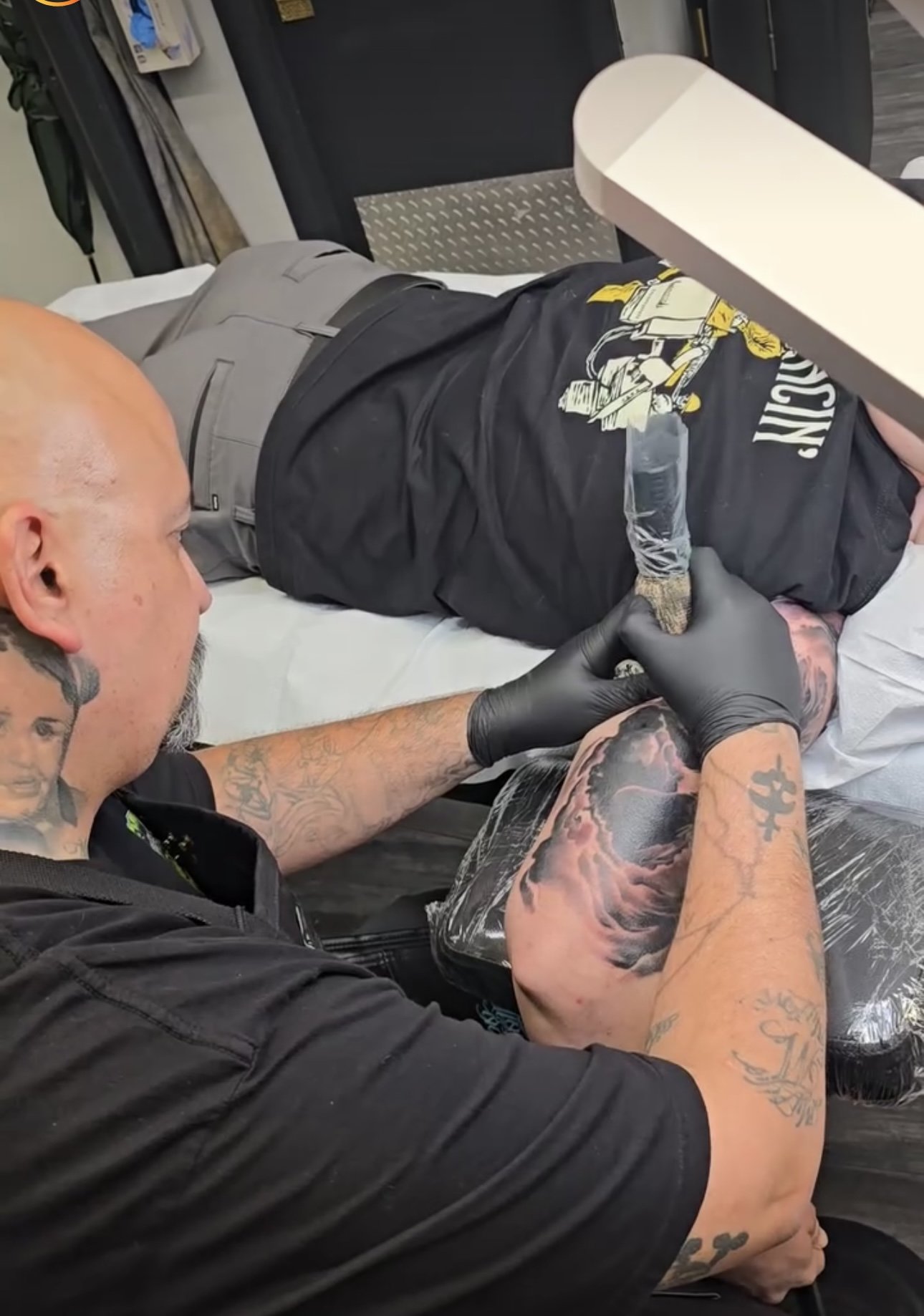 Tattoo artist creating a tattoo on a client's arm in a tattoo shop. The artist is wearing black gloves and has tattoos on his arms. The client is lying on a tattoo bed, and the focus is on the tattooing process.