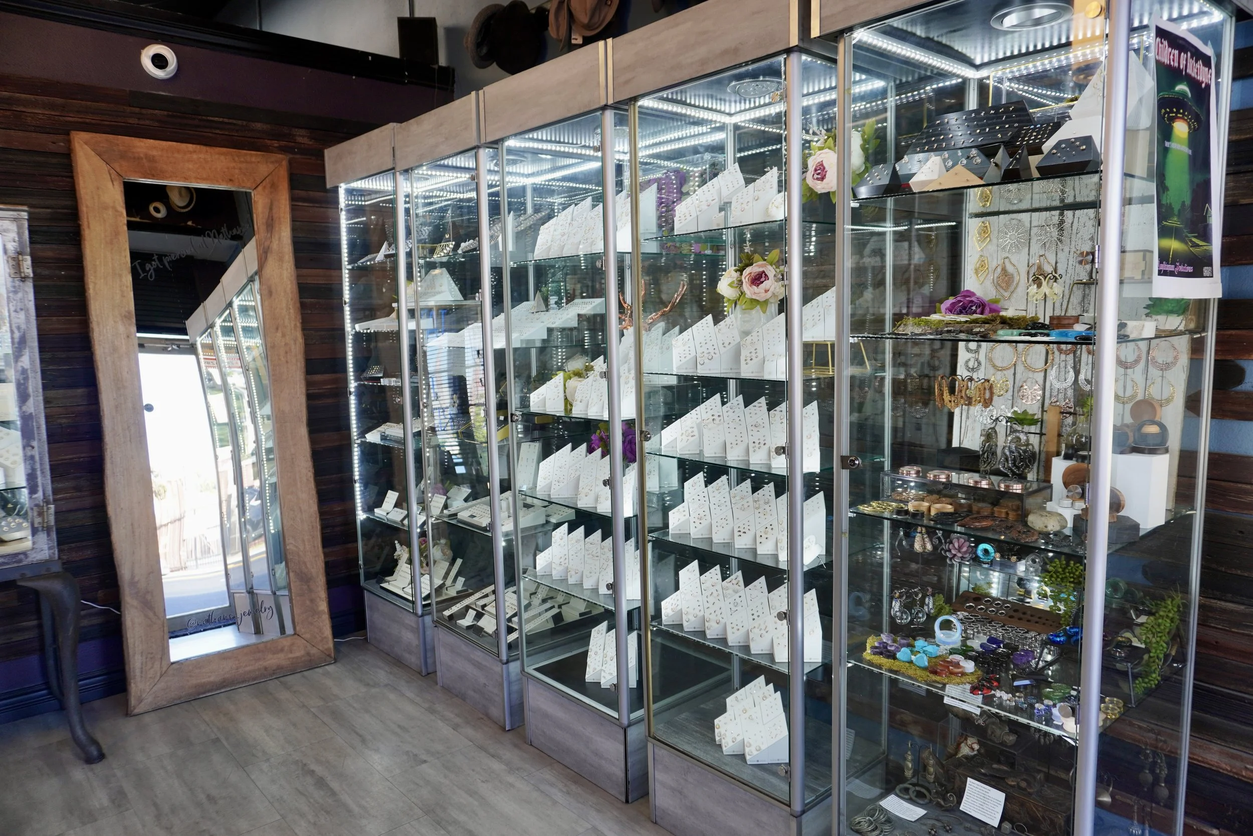 A display case filled with jewelry, including earrings, rings, bracelets, and necklaces, decorated with flowers and decorative items. To the left, a large mirror with a wooden frame leans against a dark wood-paneled wall in a retail store.