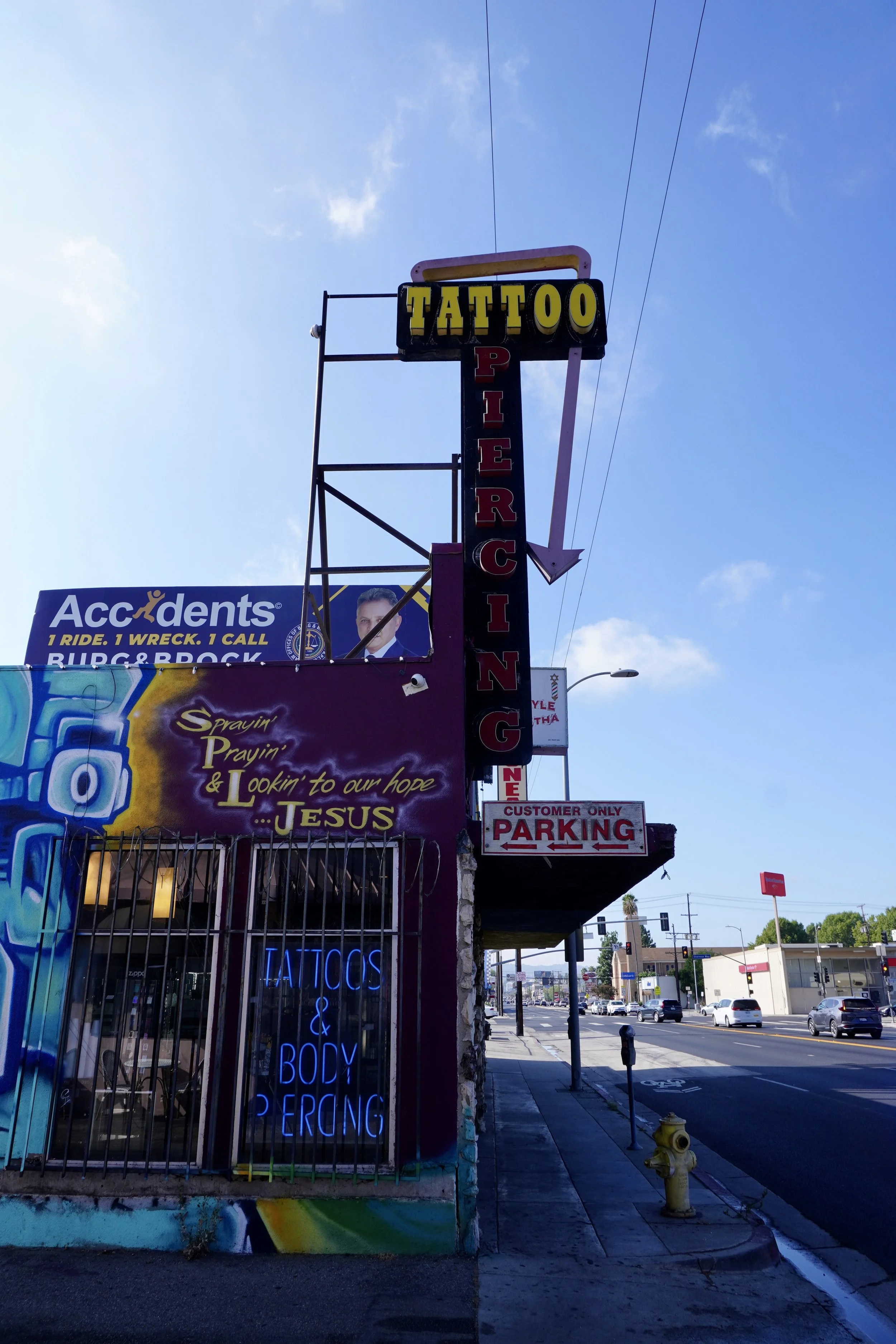 Street view of a tattoo and body piercing shop with a neon sign, a urine billboard, and a parking sign, under a partly cloudy sky.