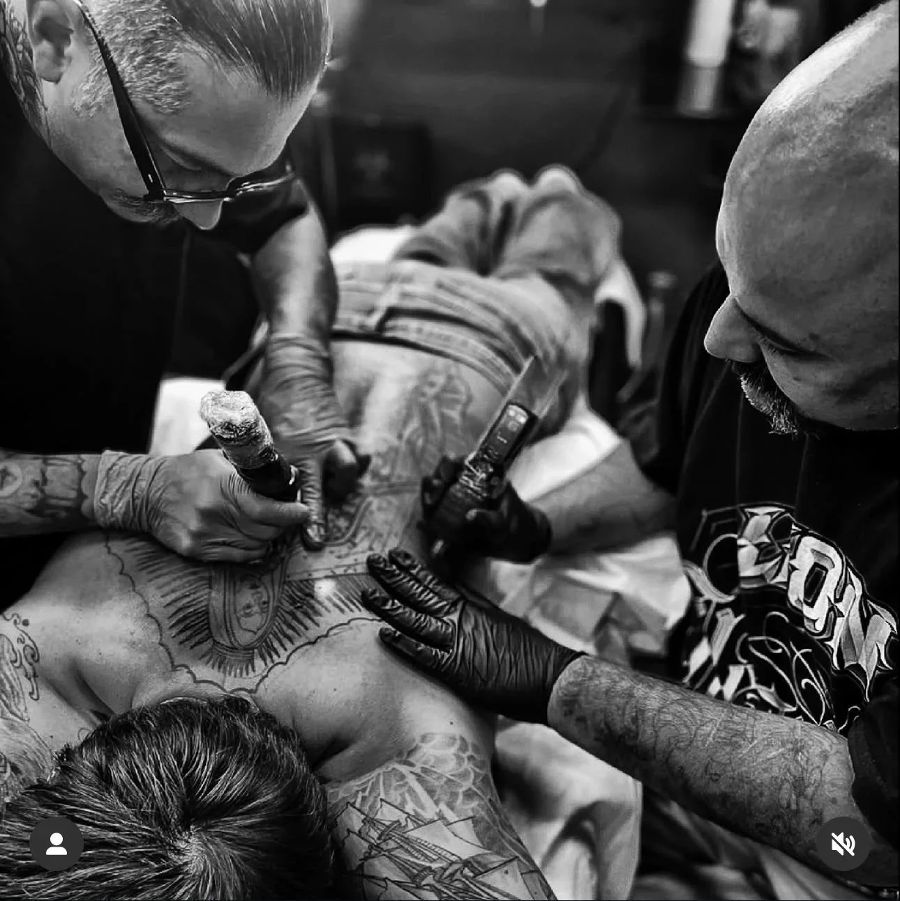 A tattoo artist is creating a tattoo on a person's back while another person assists, both wearing black gloves. The scene is in black and white.