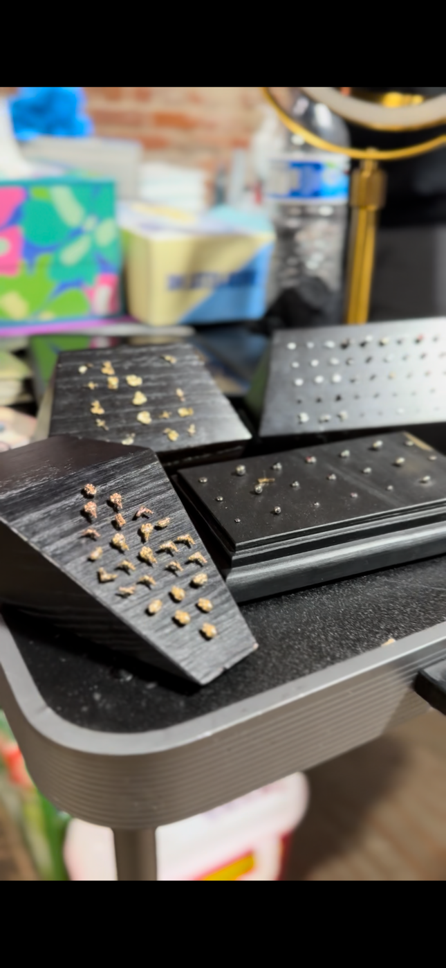 Metal jewelry or small metal parts with different textures and shapes, some arranged in lines or patterns, on black trays placed on a gray cart, in a workshop or industrial setting.