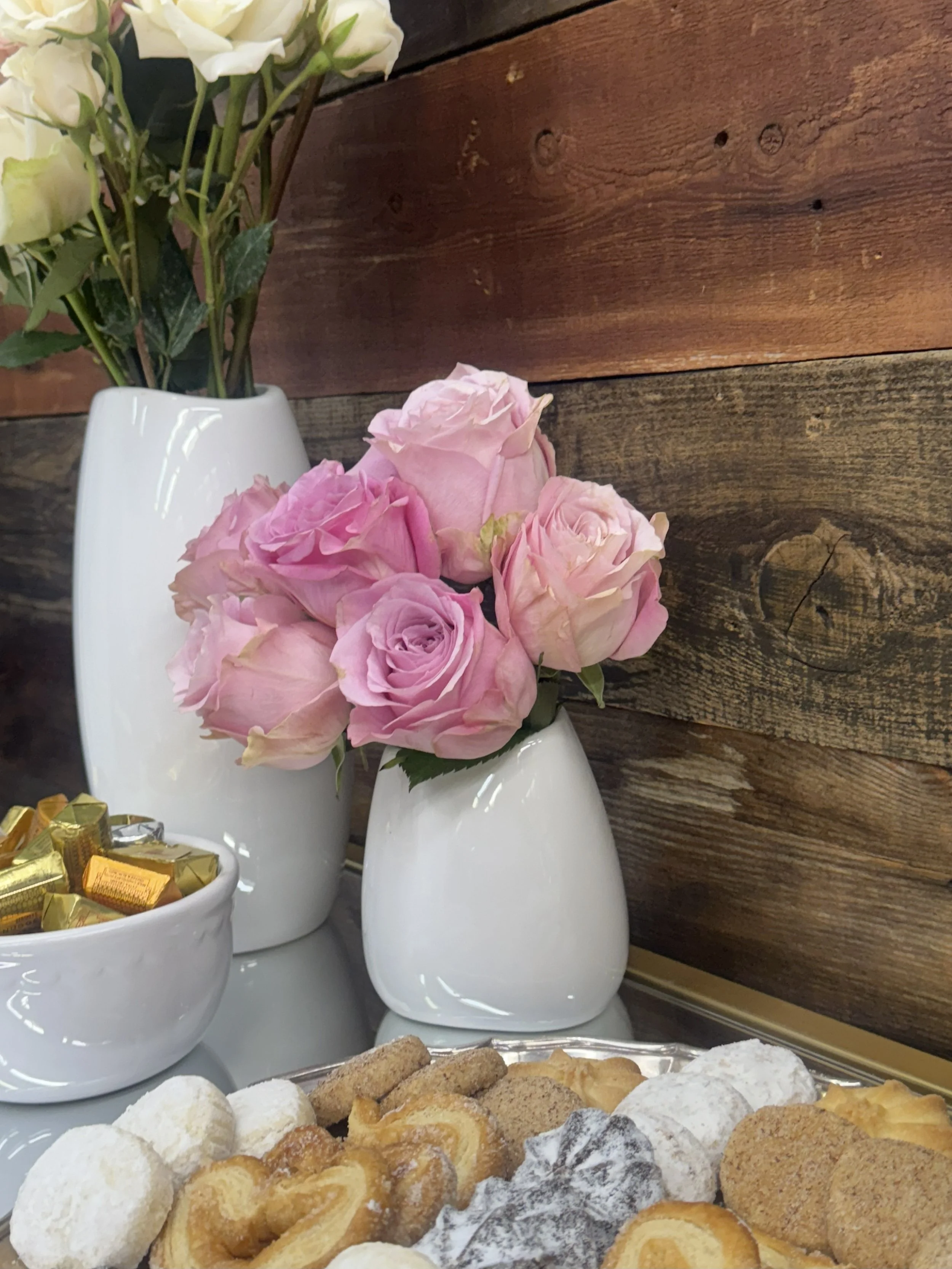 A table arrangement with a white vase containing cream-colored roses and a wall in the background made of dark wooden planks. In the foreground, there are various cookies and pastries on a tray.