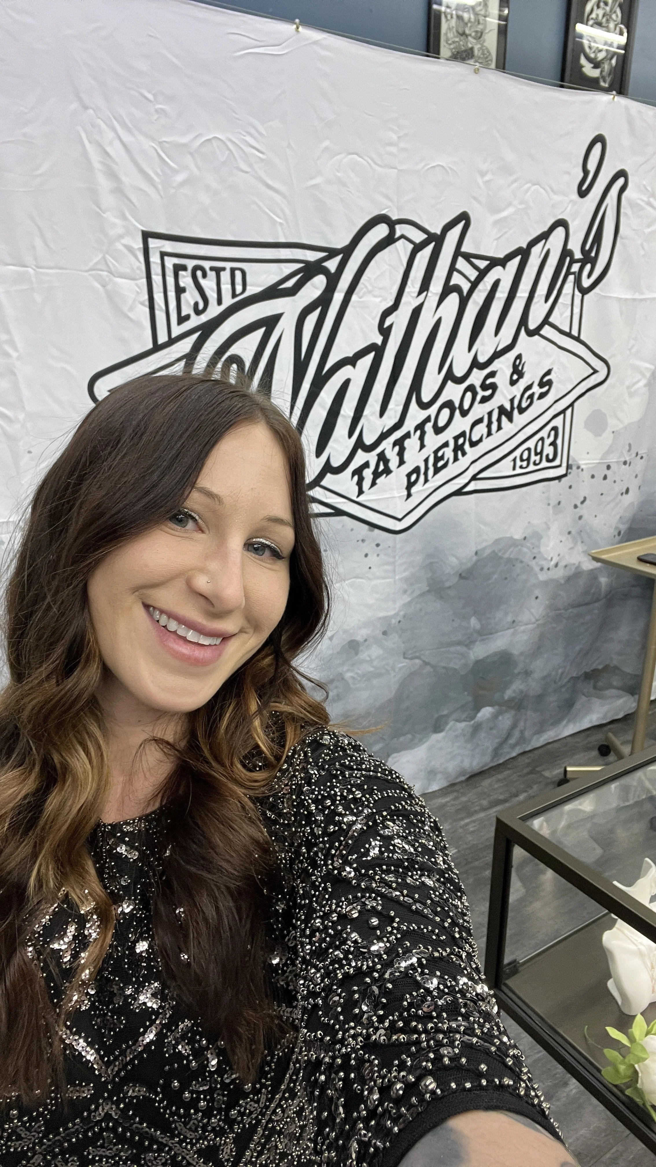A woman with long wavy hair smiling for a selfie in front of a tattoo, tattoos and piercings sign in a tattoo parlor.