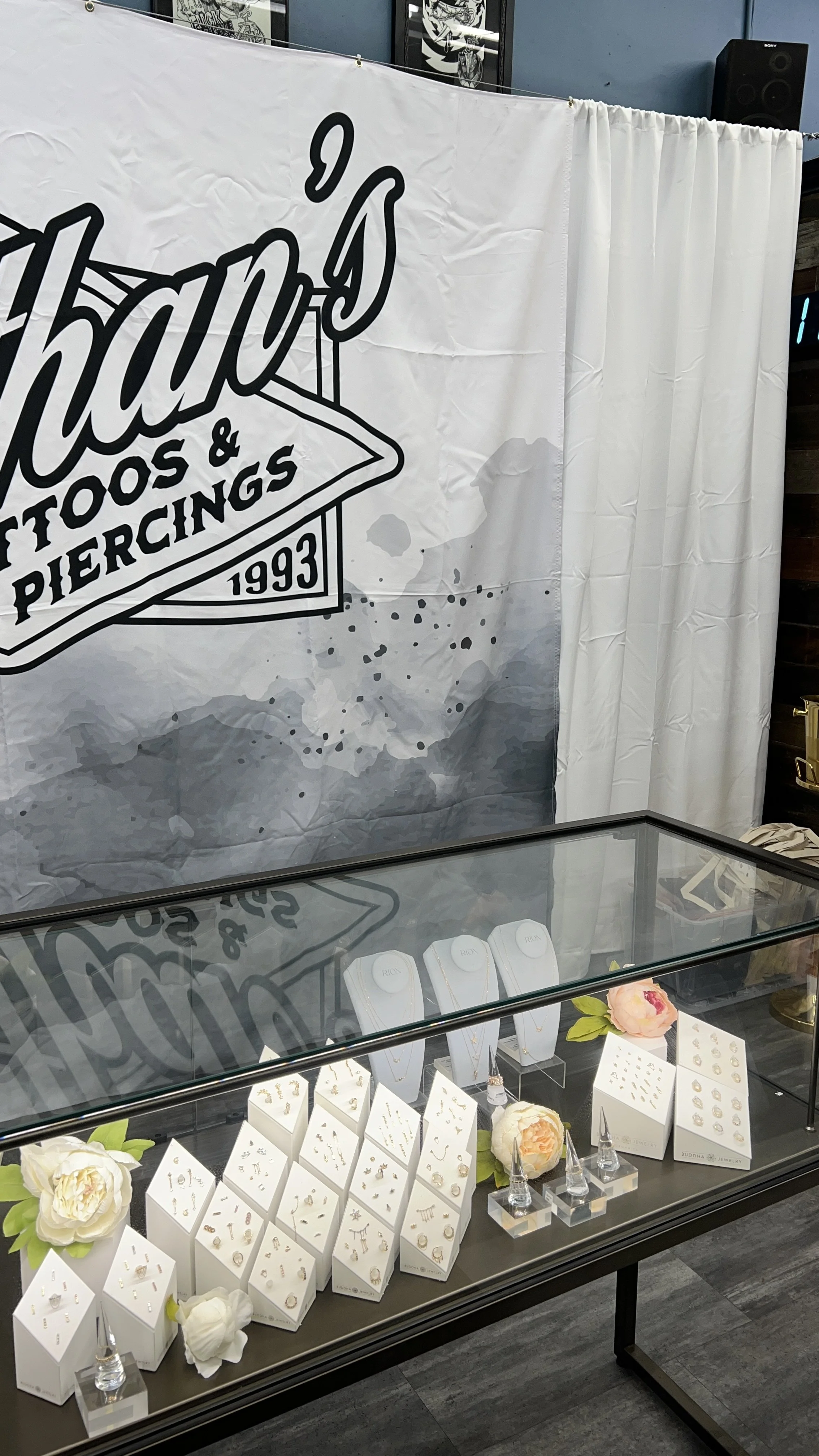 Display case with jewelry, including rings and earrings, decorated with flowers. Behind it is a large banner with a tattoo and piercing shop logo, and a white curtain.