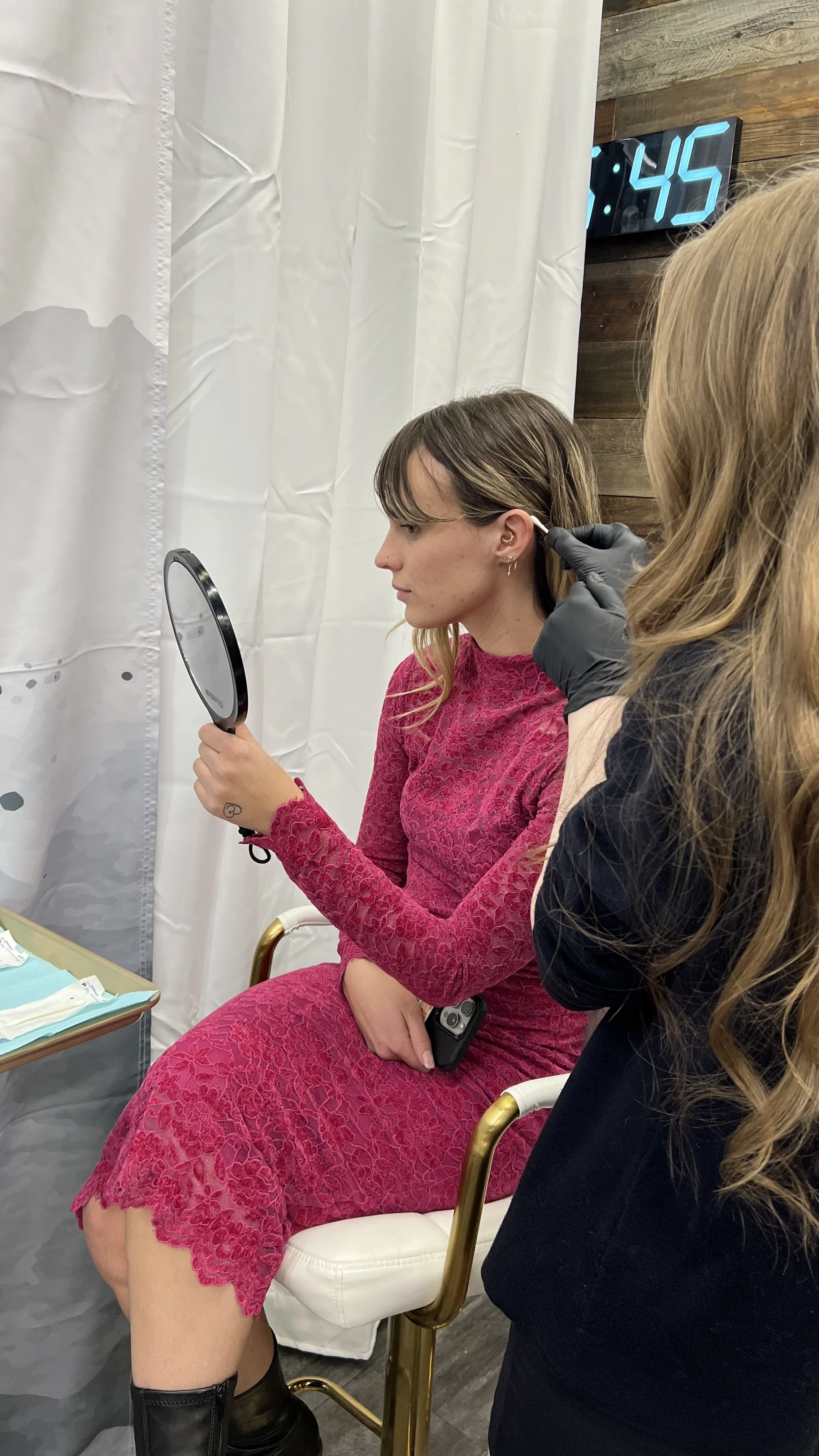 A woman in a pink lace dress sitting in a chair receiving an ear piercing from a person with black gloves. She is holding a mirror to examine her new piercing. There is a table with supplies beside her, a clock showing 7:45, and white curtains in the