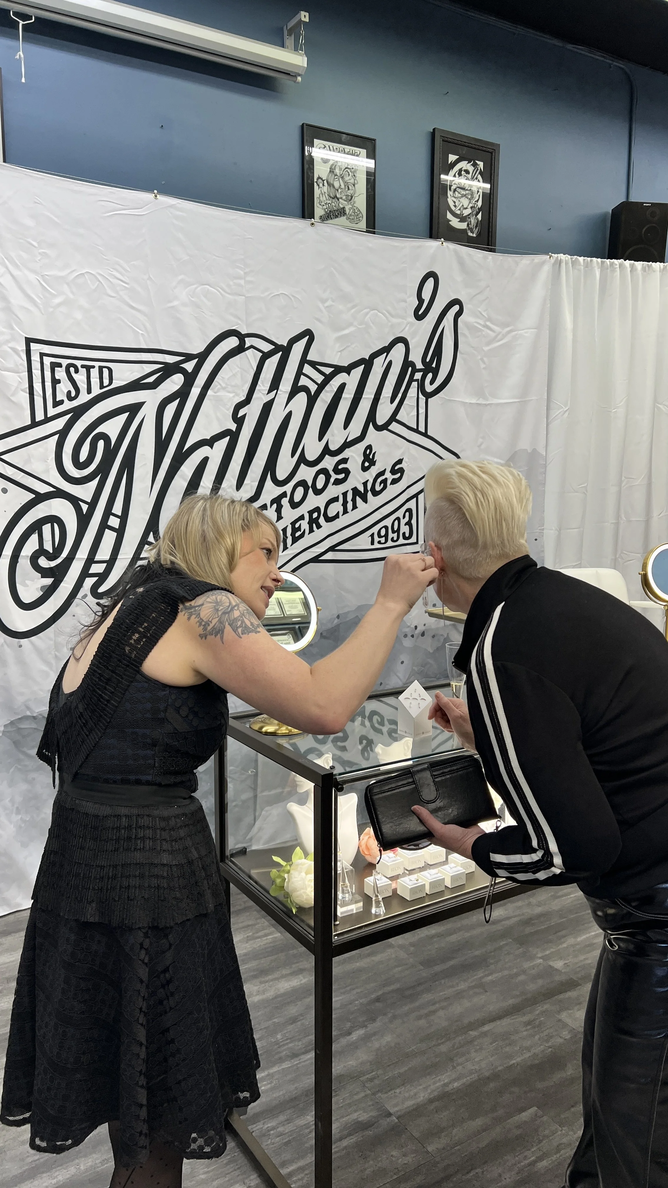A woman with blonde hair and tattoos on her arm is showing a piece of jewelry to a blonde man with a short hairstyle, who is holding a black wallet and a phone. They are standing in front of a display case with jewelry, inside a store with a banner t
