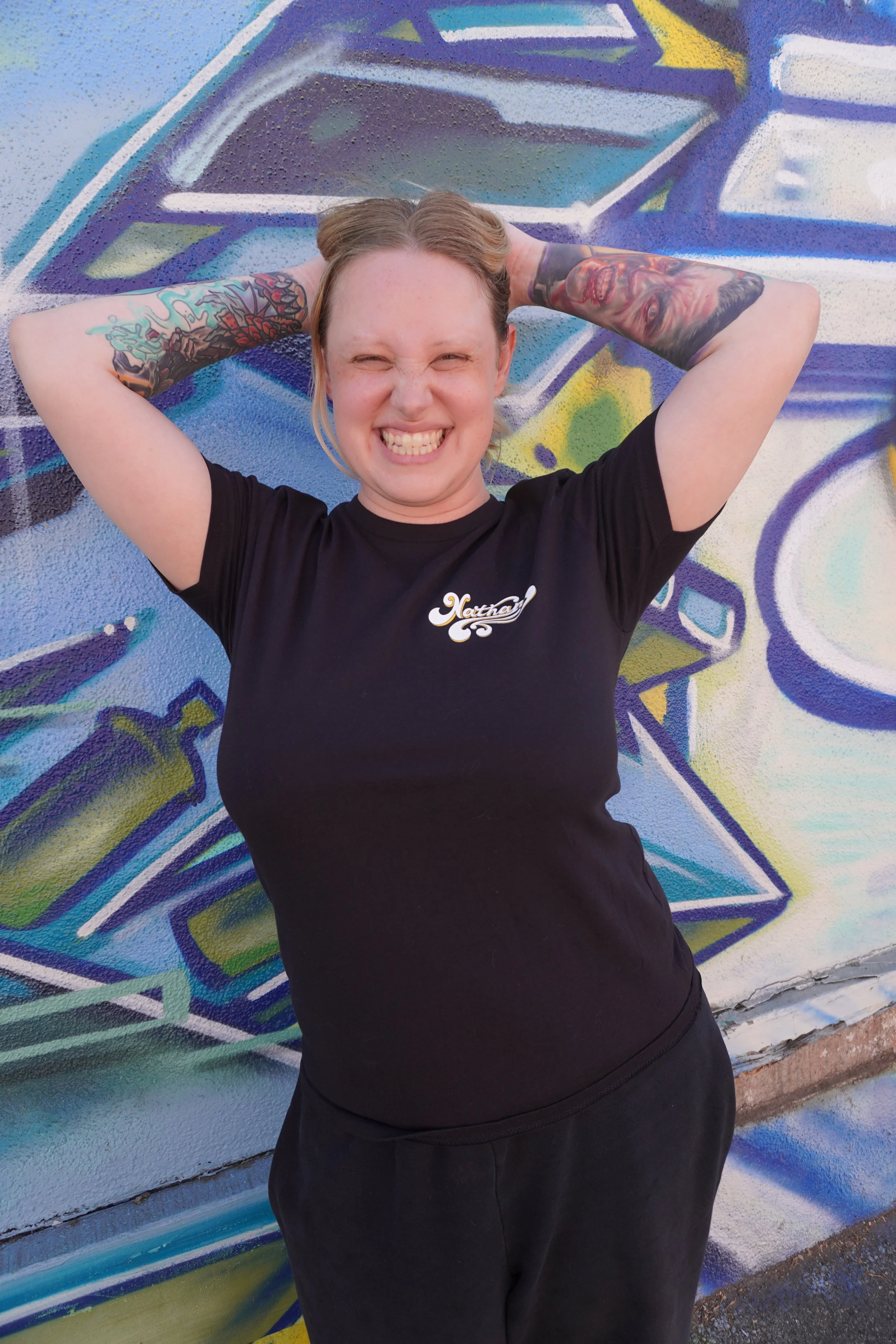 Young woman smiling with eyes closed, arms behind head, standing in front of a colorful graffiti mural.