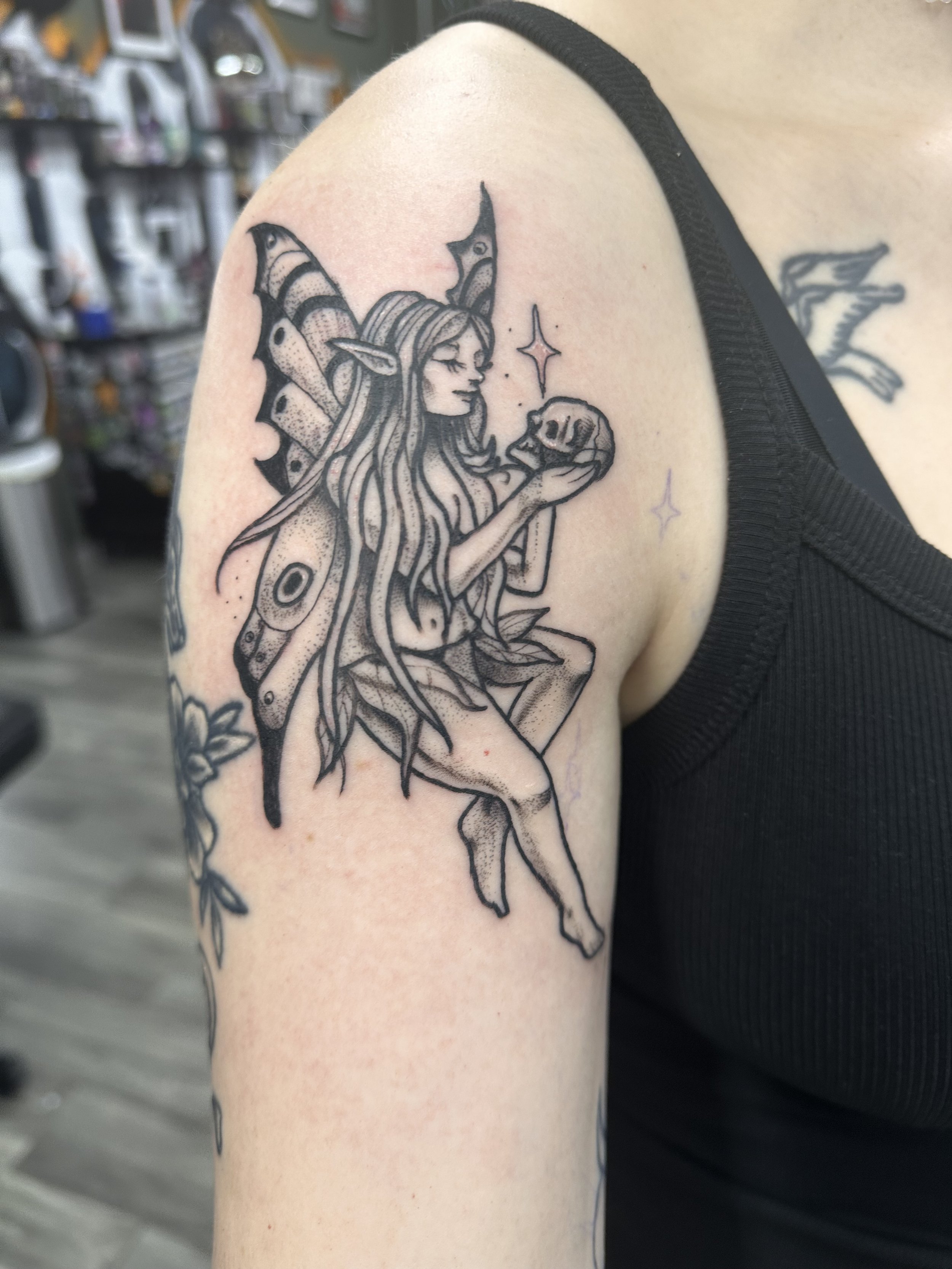 Tattoo of a fairy with butterfly wings holding a skull, with star accents, on a person's shoulder.
