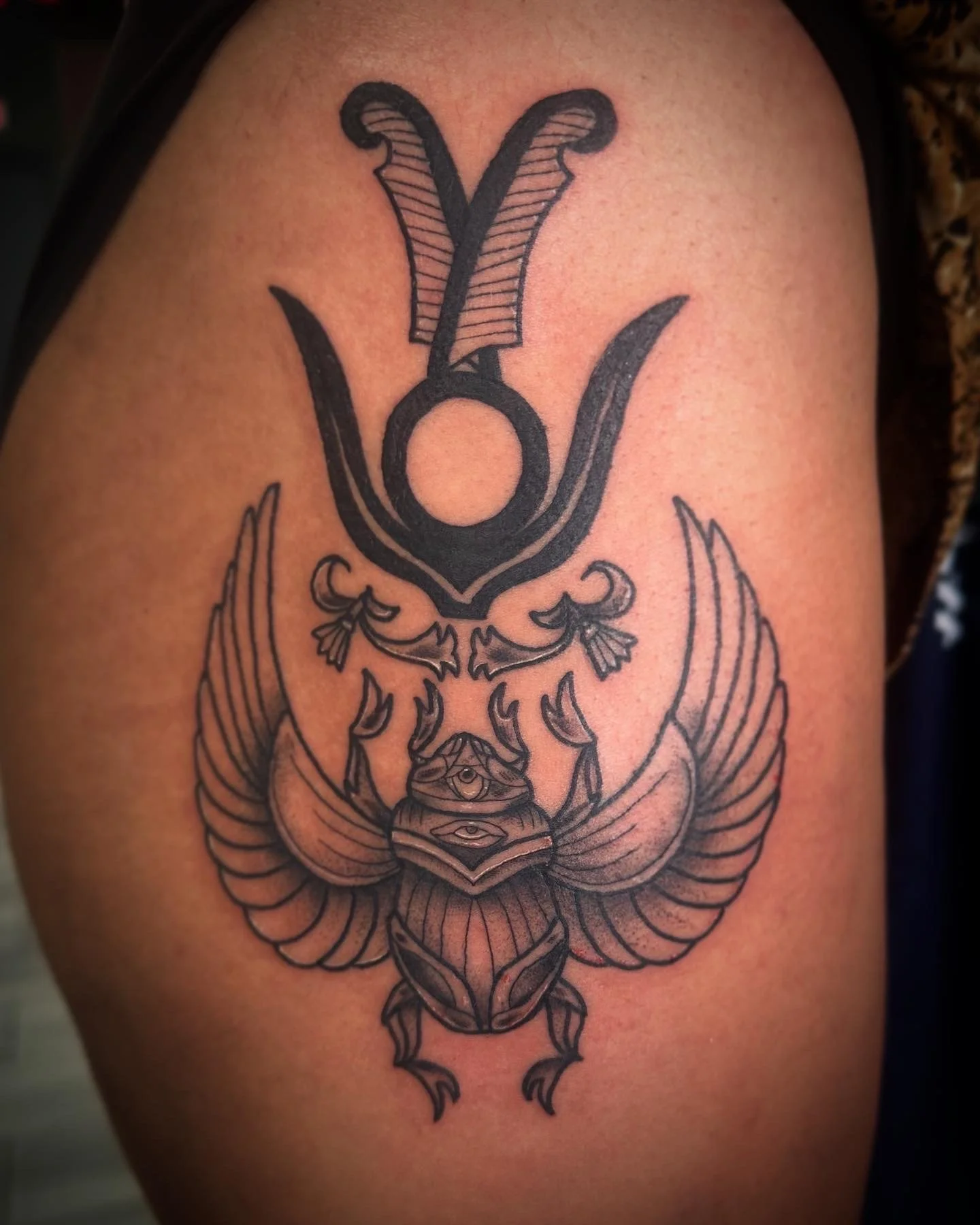 Tattoo of a scarab beetle with detailed wings and an eye on its back, surrounded by decorative elements and patterns, on a person's upper arm.