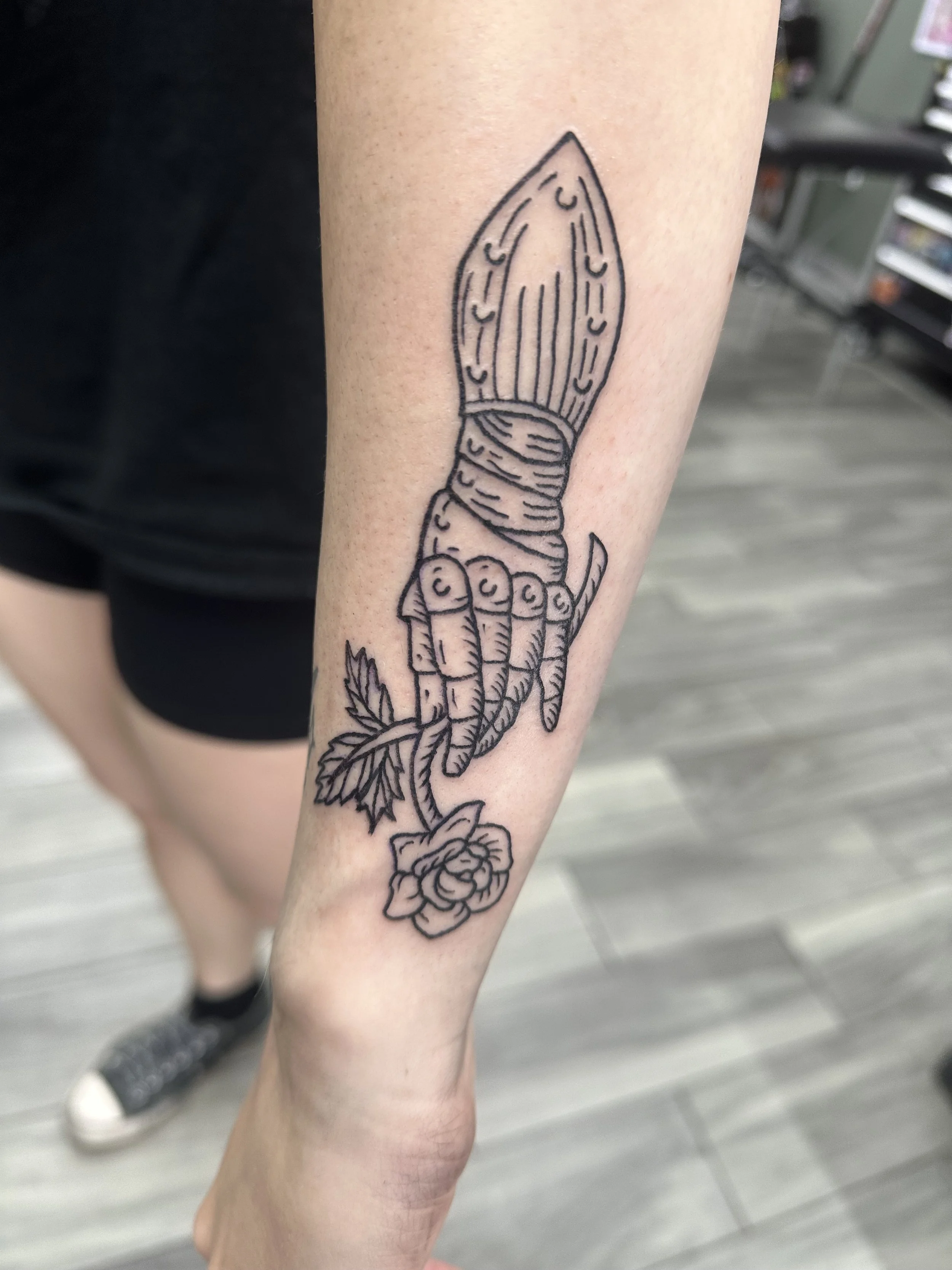 A tattoo of a hand gripping a large knife with a rose at the bottom, inked on a person's forearm.