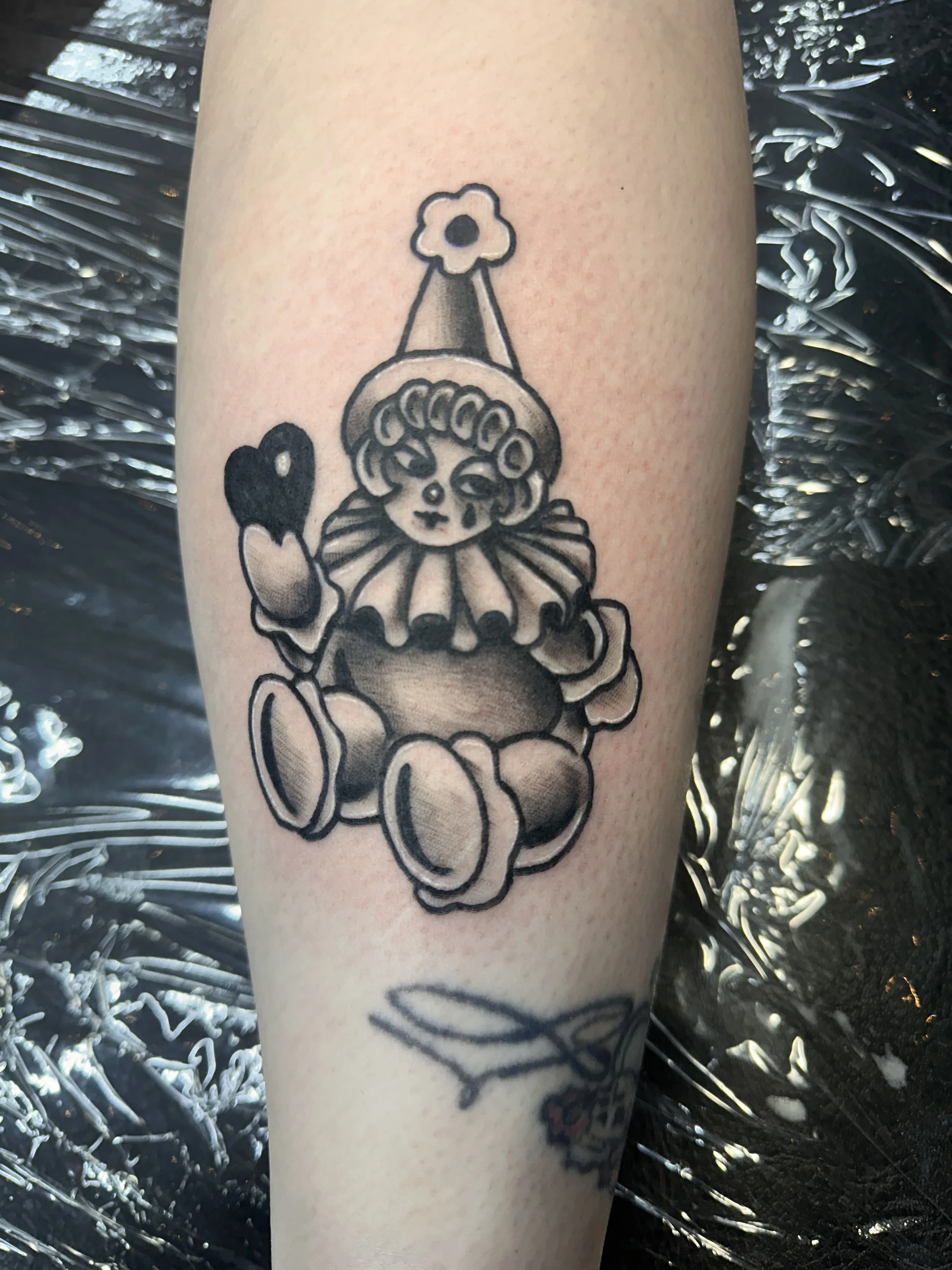 Tattoo of a clown with a flower cone hat holding a heart, on a person's forearm.