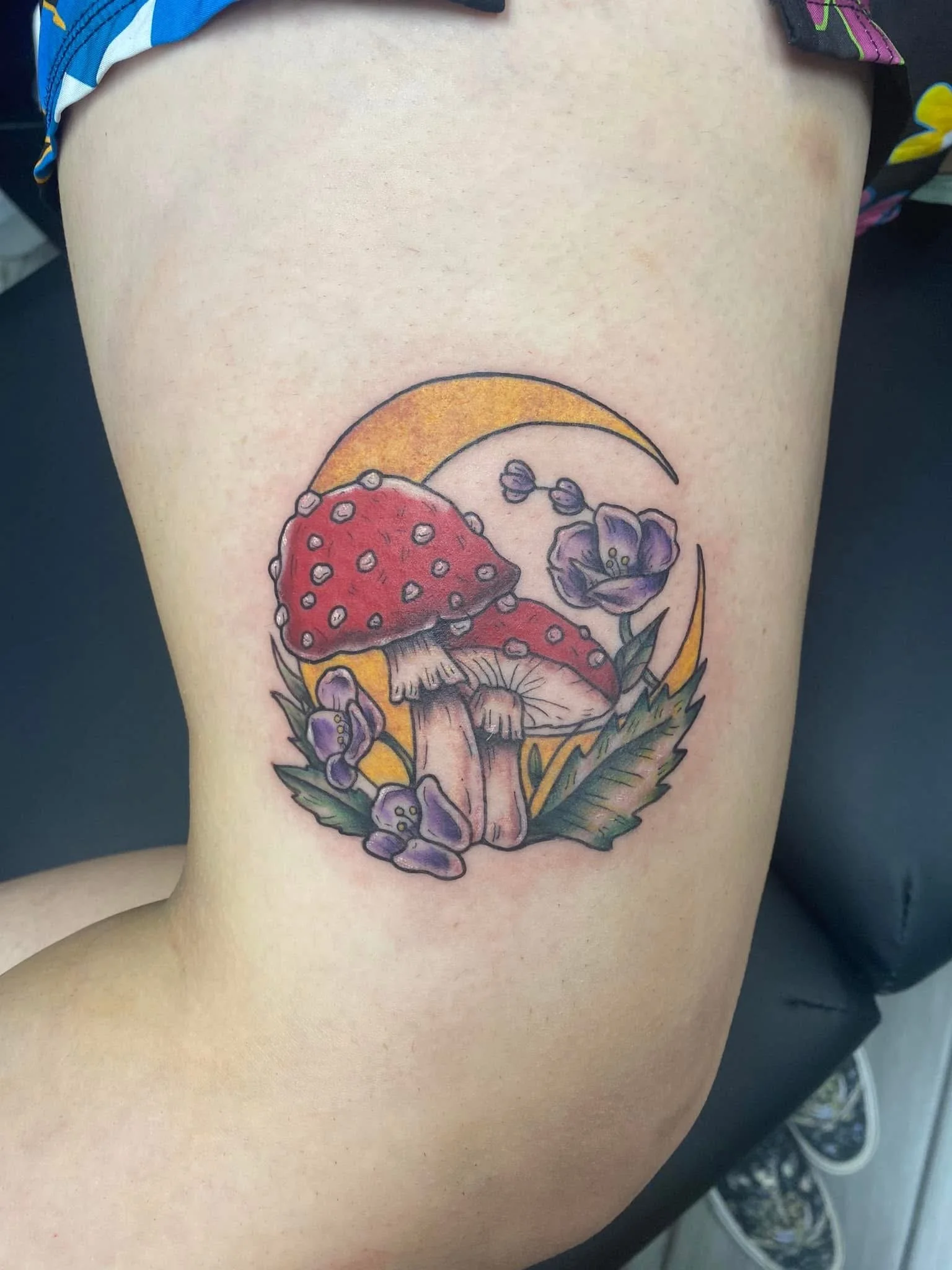 A colorful tattoo on a person's side featuring mushrooms, purple flowers, leaves, and a yellow crescent shape.