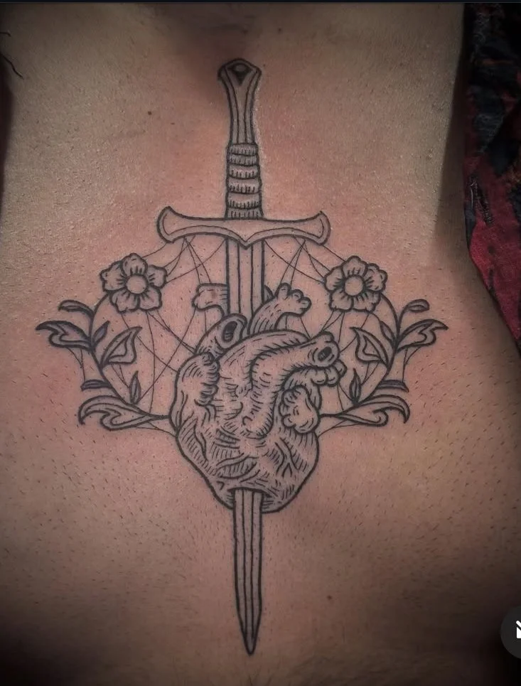 Tattoo of a heart with a sword piercing through it, surrounded by flowers and a web pattern behind it