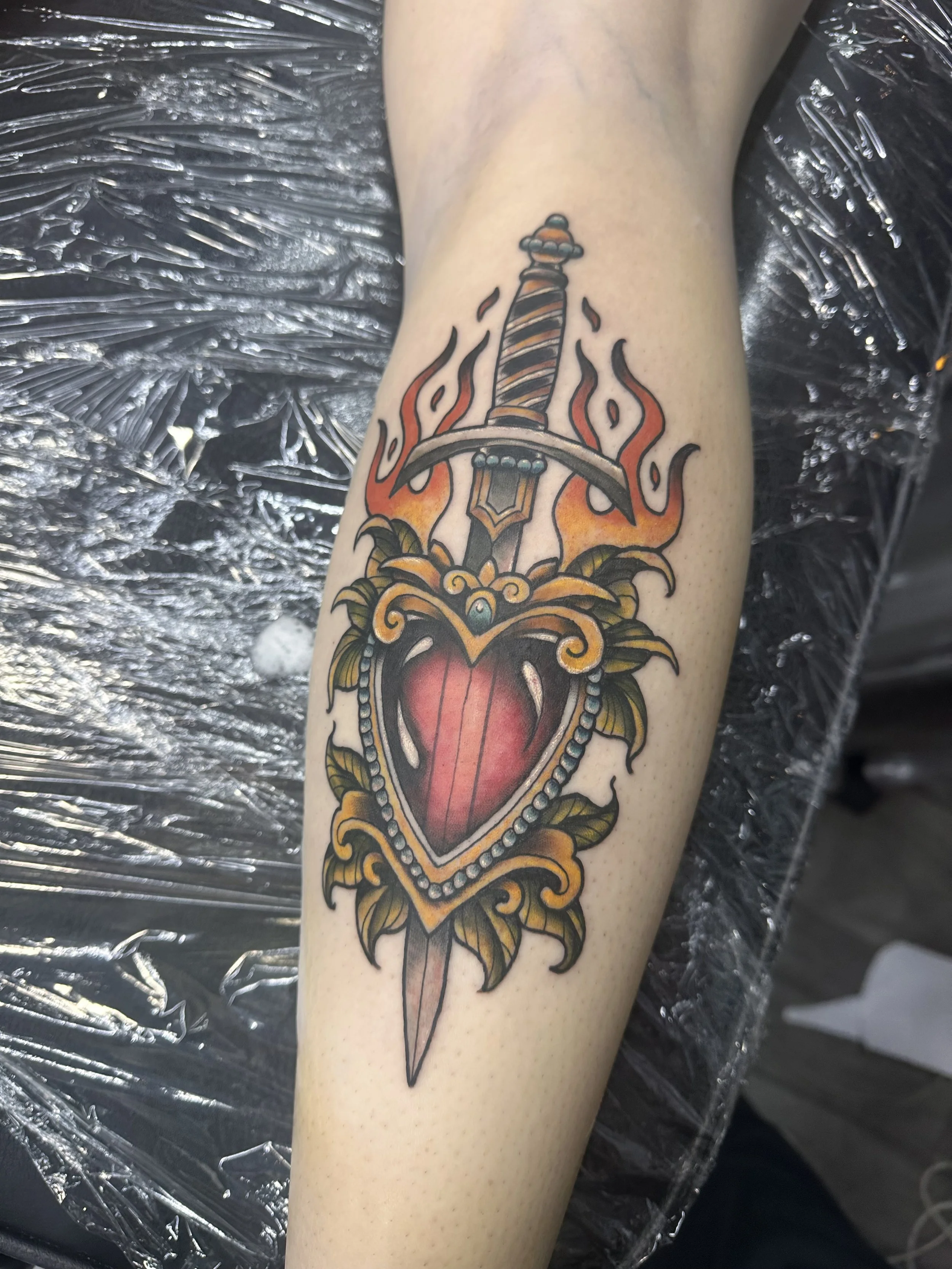 A tattoo of a sword with a heart-shaped blade, surrounded by flames and decorative elements, on a person's forearm.