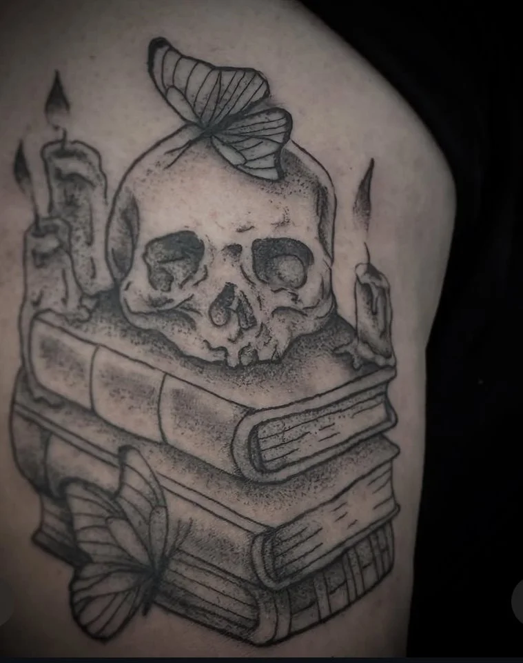 Tattoo of a skull on top of stacked books, with a butterfly perched on the skull and another near the bottom book, and candles around the skull.