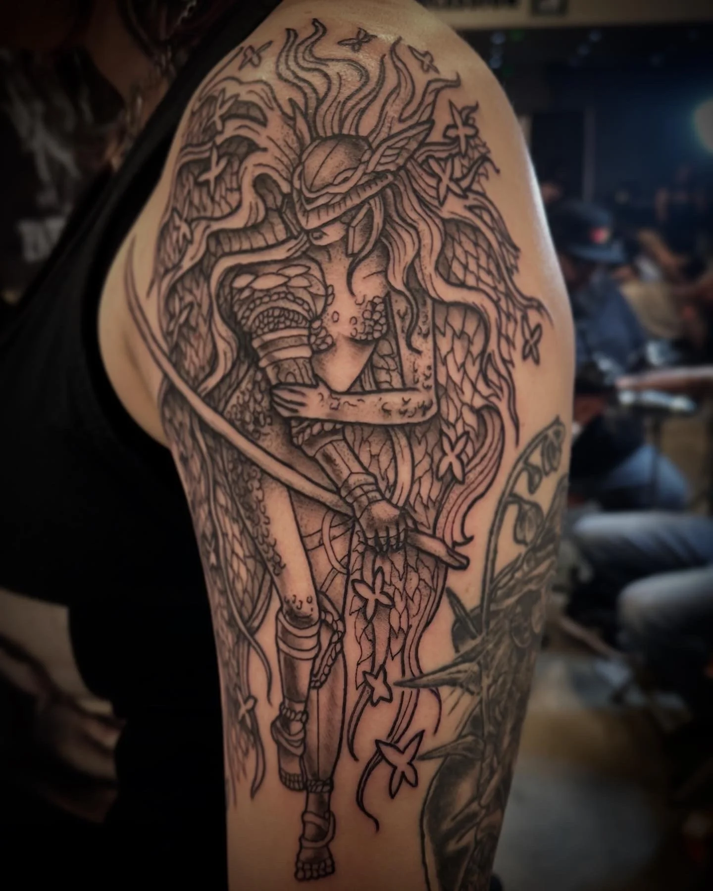 Blackline tattoo of a woman with flowing hair, wearing a wide-brimmed hat, holding a sword, surrounded by butterflies and floral elements, on a person's upper arm.
