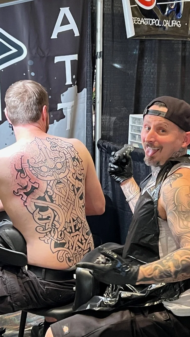 A tattoo artist with tattoos on his arms and wearing a backward cap is posing happily with his tongue out, holding a tattoo machine, beside a man with a large back tattoo that is still in progress at a tattoo convention.