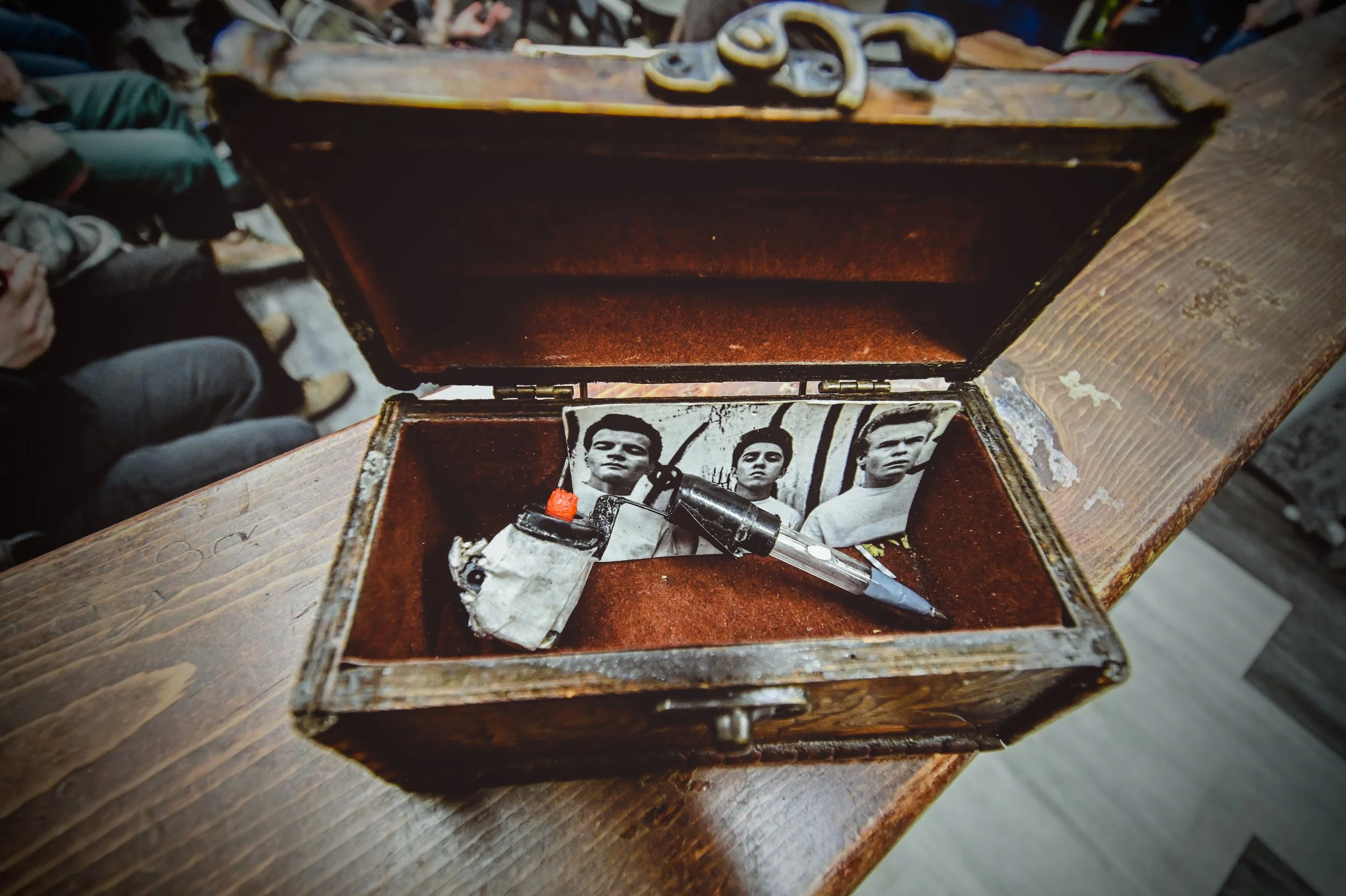 An open wooden box containing a black spray paint can, a small white paint tube, and a black spray paint nozzle. Inside the box are black and white photographs of young men with serious expressions.
