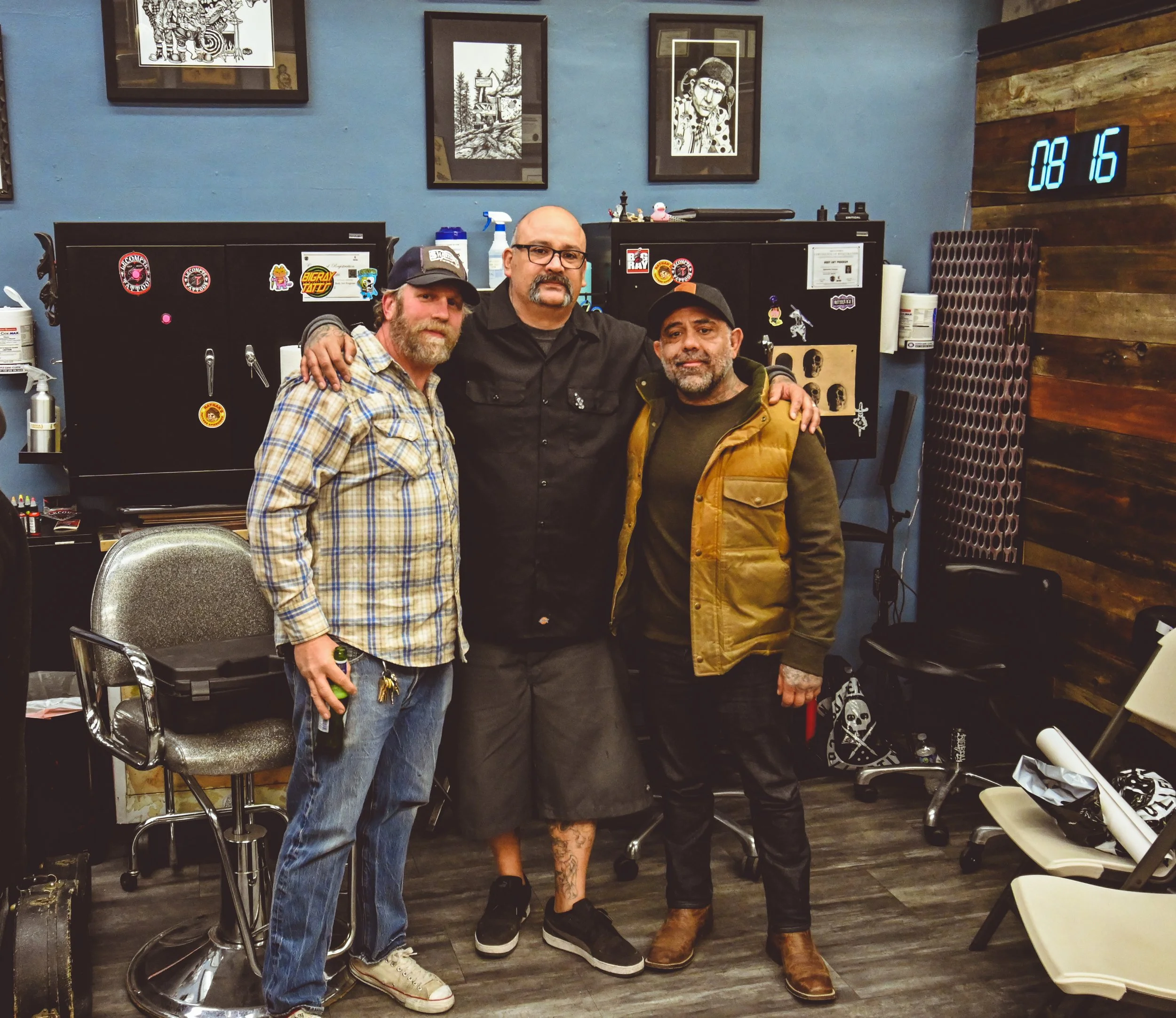 Three men standing together in a tattoo shop, with black lockers decorated with stickers behind them, a blue wall with framed artwork, and a digital clock displaying 08:16 on a wooden paneled wall.