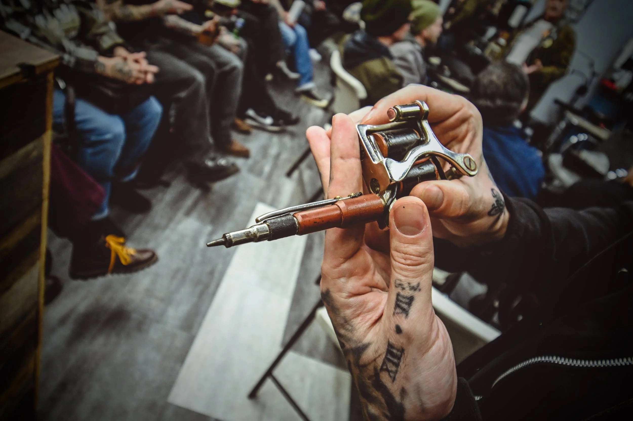 Close-up of a tattooed hand holding a revolver-style tattoo machine, with a row of seated people in a workshop or classroom setting in the background.