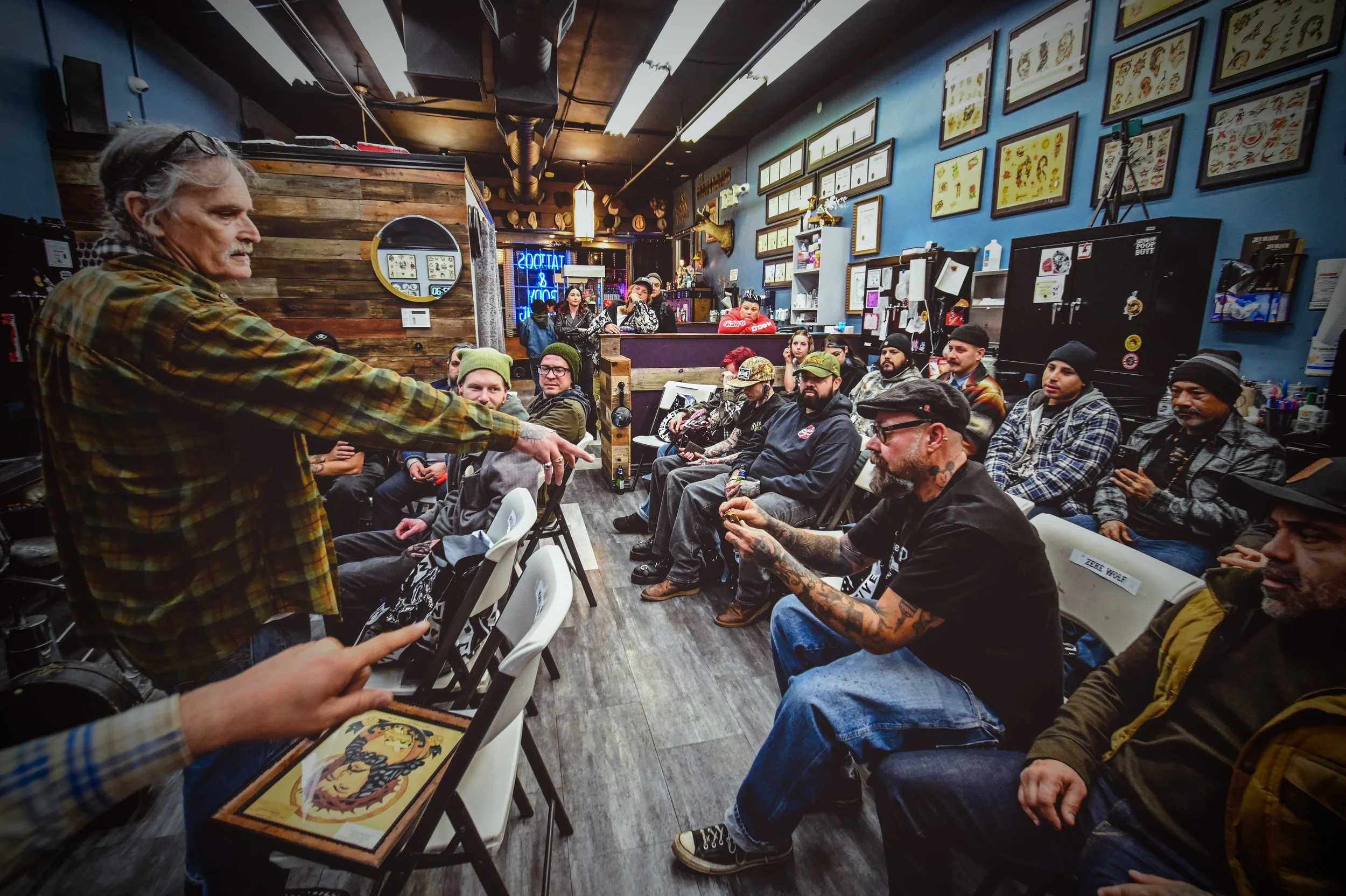A man with gray hair and glasses, wearing a yellow and brown plaid shirt, points towards a group of seated people inside a tattoo shop. The room has a blue wall decorated with framed artwork and certificates, a wooden partition, and a neon sign that 