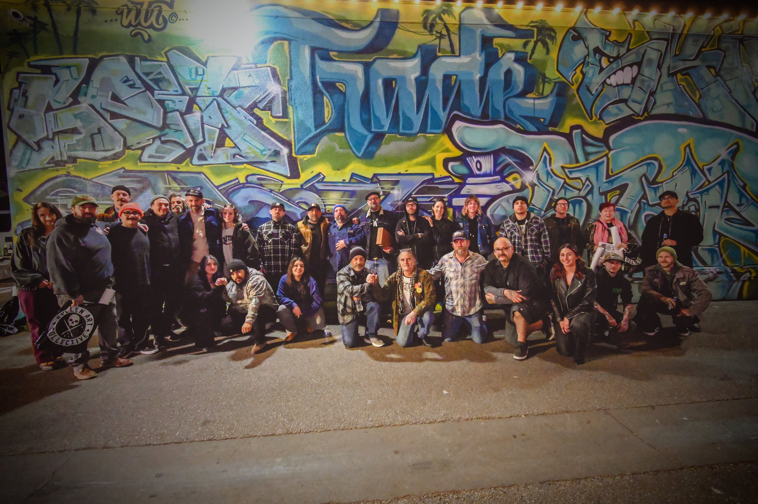 A group of people posing in front of a large colorful graffiti mural on a wall, with some of them kneeling in the front row and others standing behind.