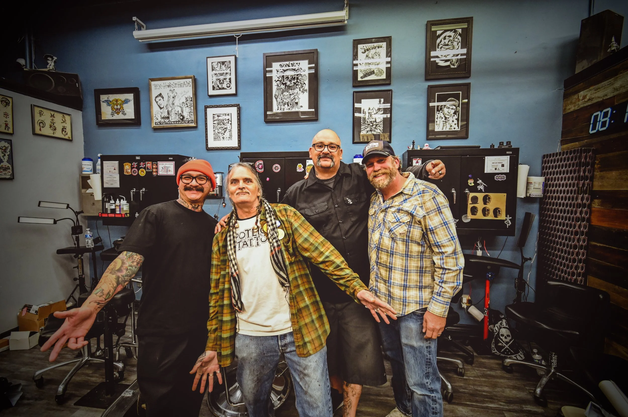 Four men standing together and smiling inside a tattoo parlor with framed artwork on the blue wall behind them.