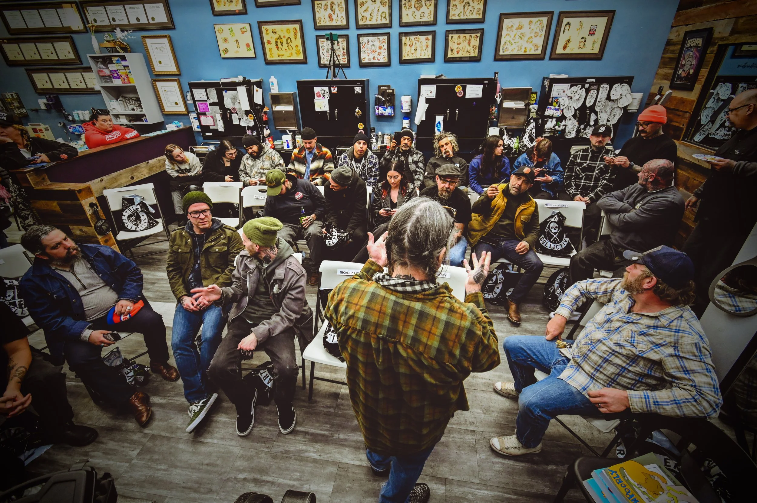 A group of people gathered in a room with blue walls and framed artwork, seated on chairs, some engaging in conversation, and a person standing at the front speaking.