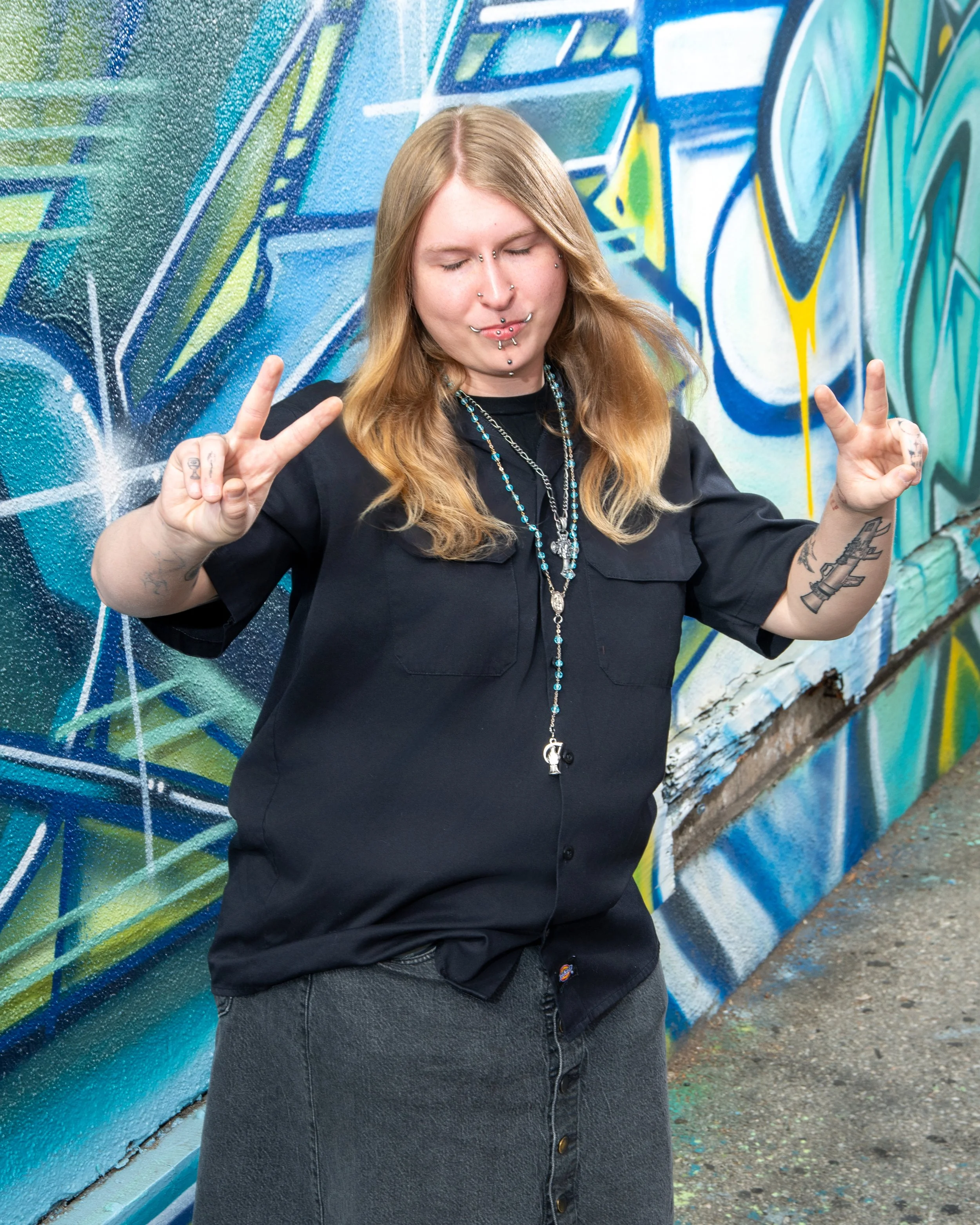 A young woman with long, wavy red hair and multiple facial piercings, including on her nose, lips, and eyebrows, posing with her eyes closed and hands raised in peace signs in front of a colorful graffiti wall.