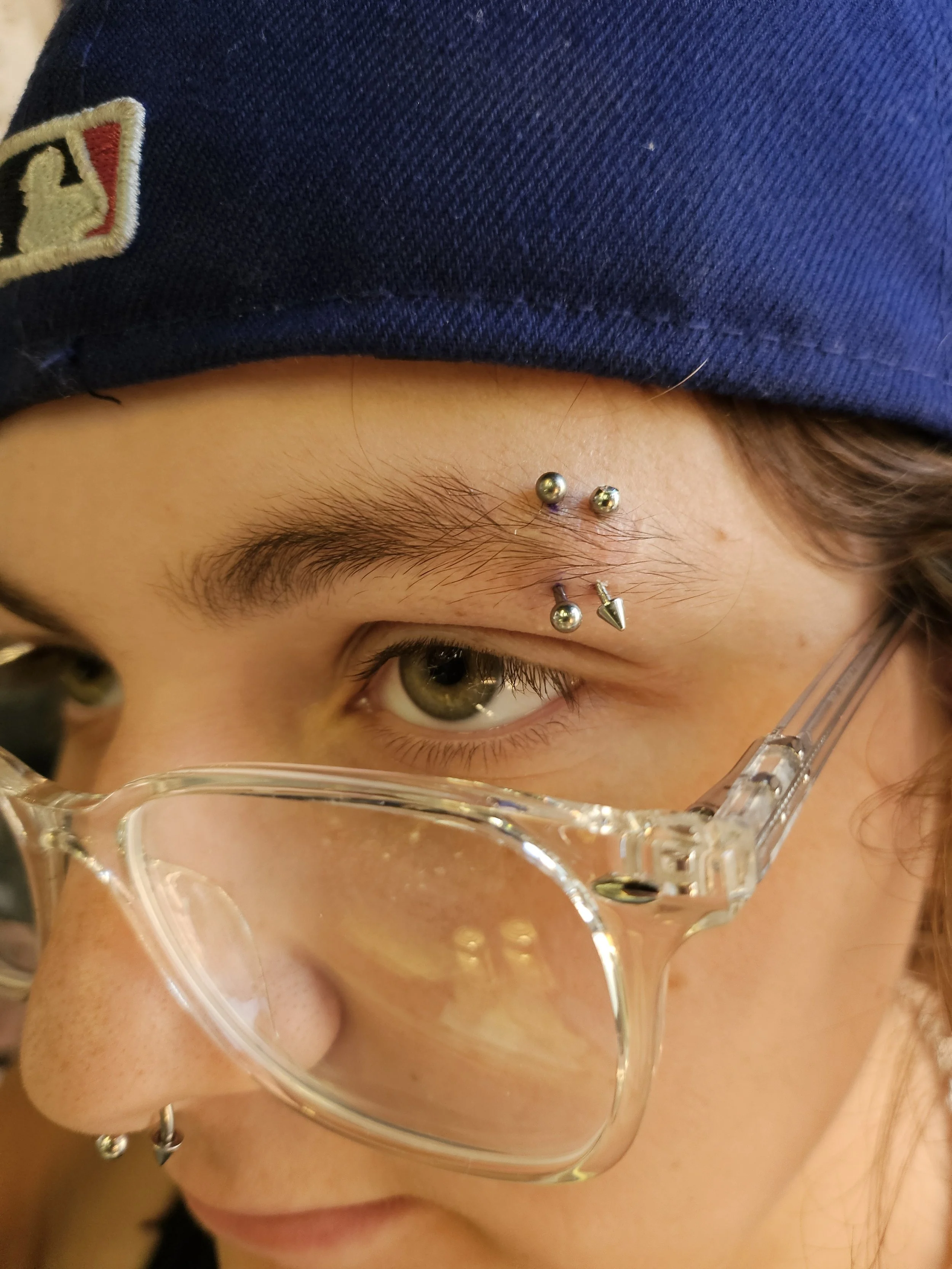 Close-up of a person's face showing eye, eyebrow, and forehead, with multiple facial piercings including a bridge piercing, a septum piercing, and a Monroe piercing, wearing clear glasses and a dark blue cap.