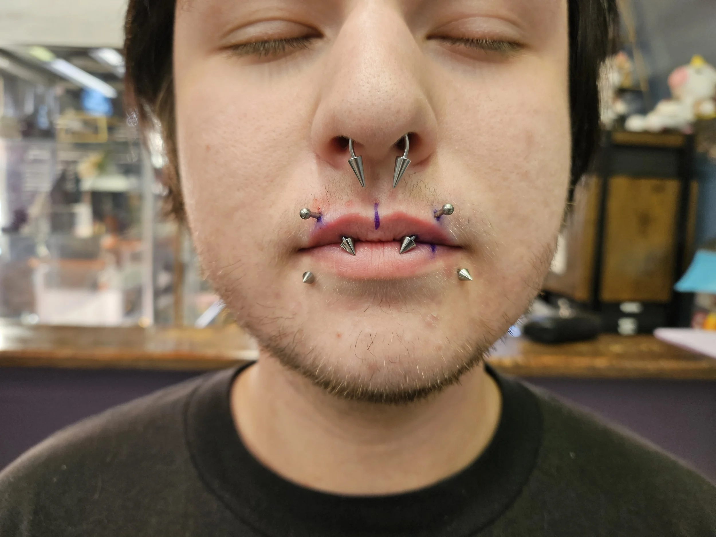A close-up of a person's face with eyes closed, showing multiple facial piercings including a septum ring with spikes, and piercings on the lips. The person has dark hair and is wearing a black shirt.