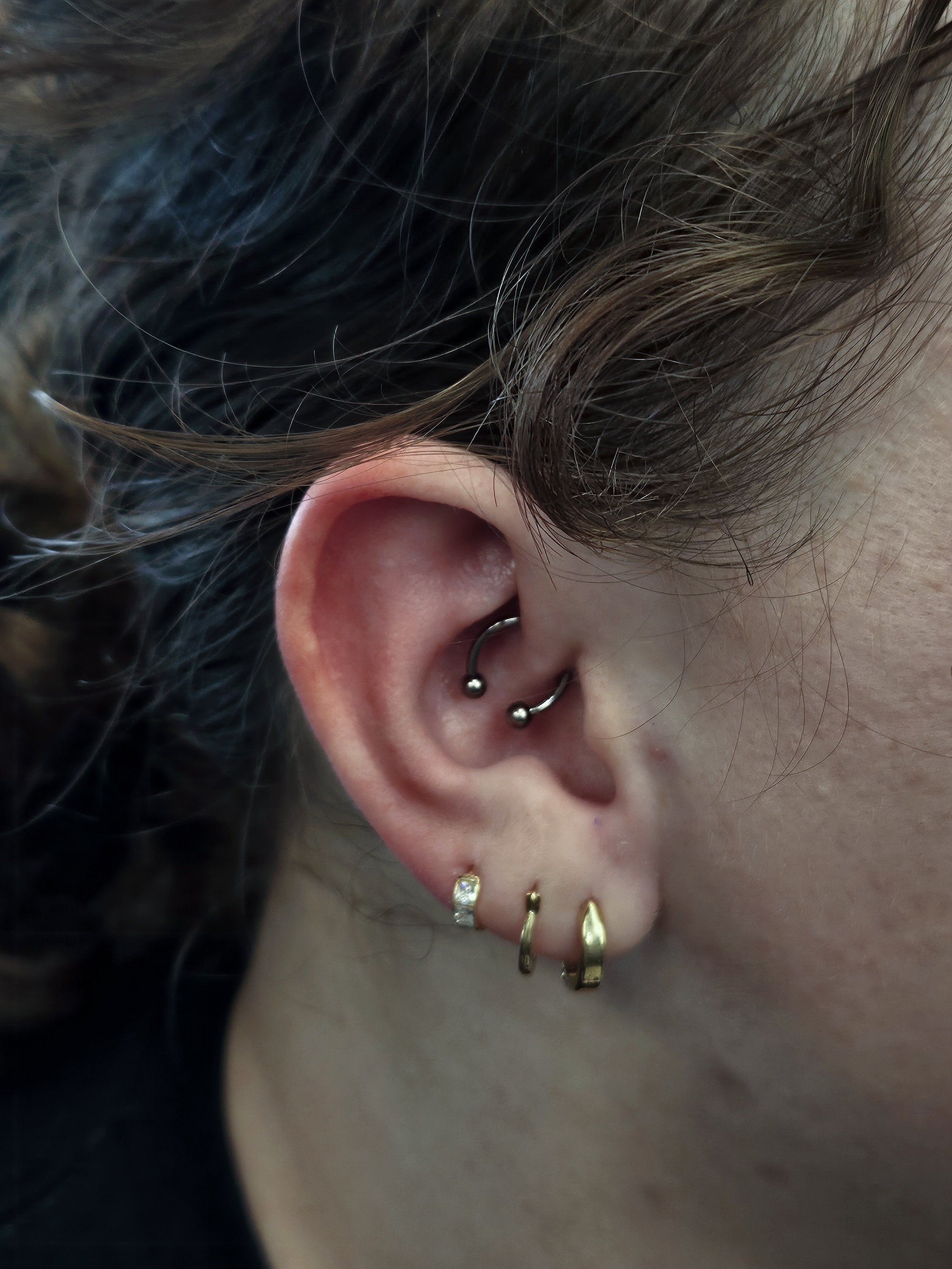 Close-up of a person's ear with multiple piercings, including two small hoop earrings and several earrings with different shapes and sizes, and brown curly hair partially covering the ear.
