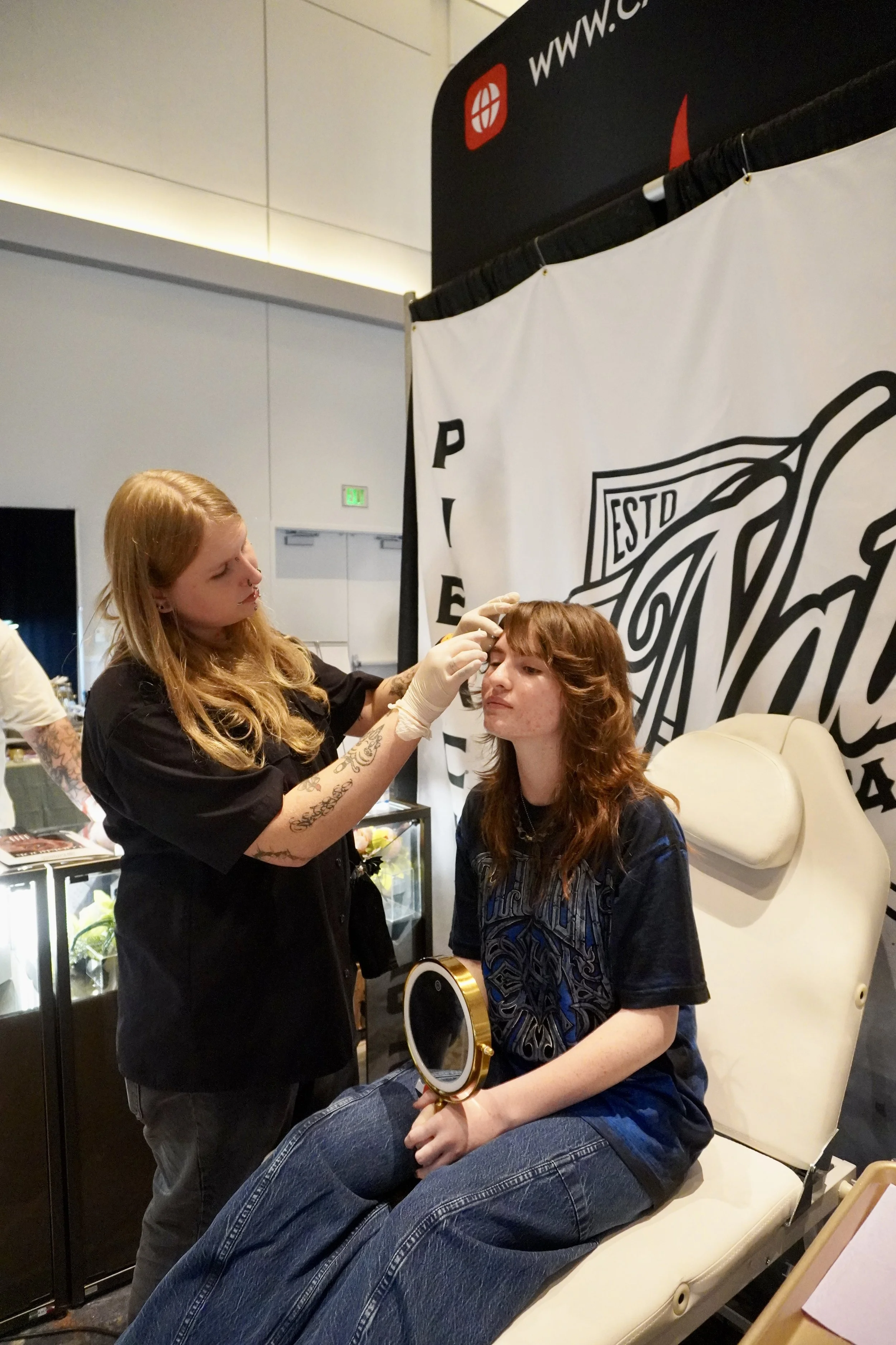 A woman sitting in a salon chair having her eyebrows shaped by a beauty technician. The technician is holding a small tool and wearing gloves. There is a large banner with black and white graphics in the background.