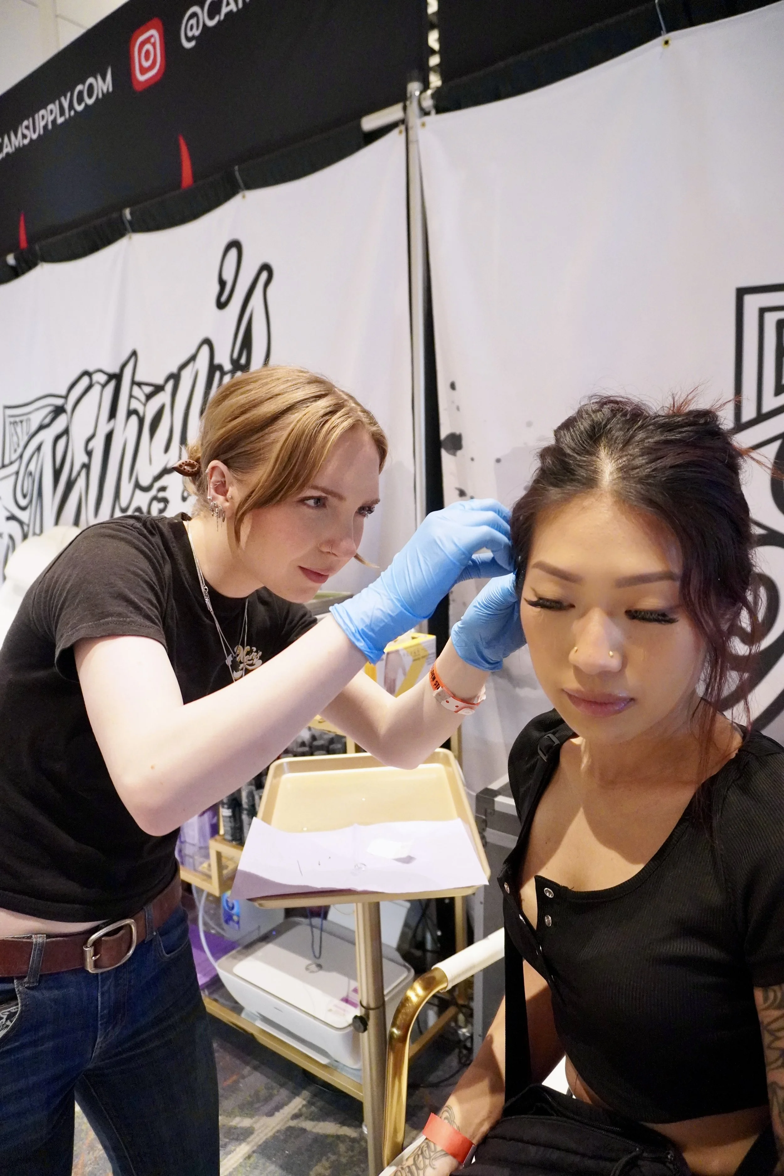 A woman with short brown hair and earrings is looking at a tattoo being applied by a tattoo artist with red hair and tattoos on her arm. The woman is sitting with her eyes closed, wearing a black outfit, and has a red wristband. The tattoo artist is 