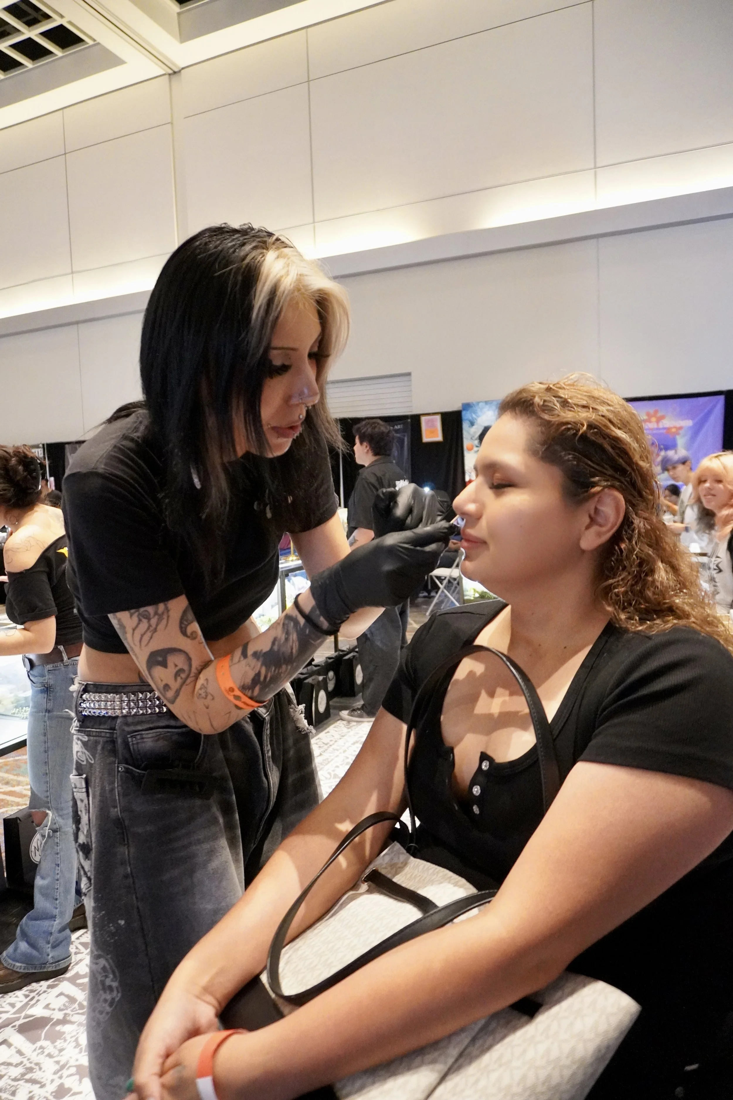 A woman with tattoos on her arms is applying makeup or tattooing on another woman with curly hair at a convention or event.