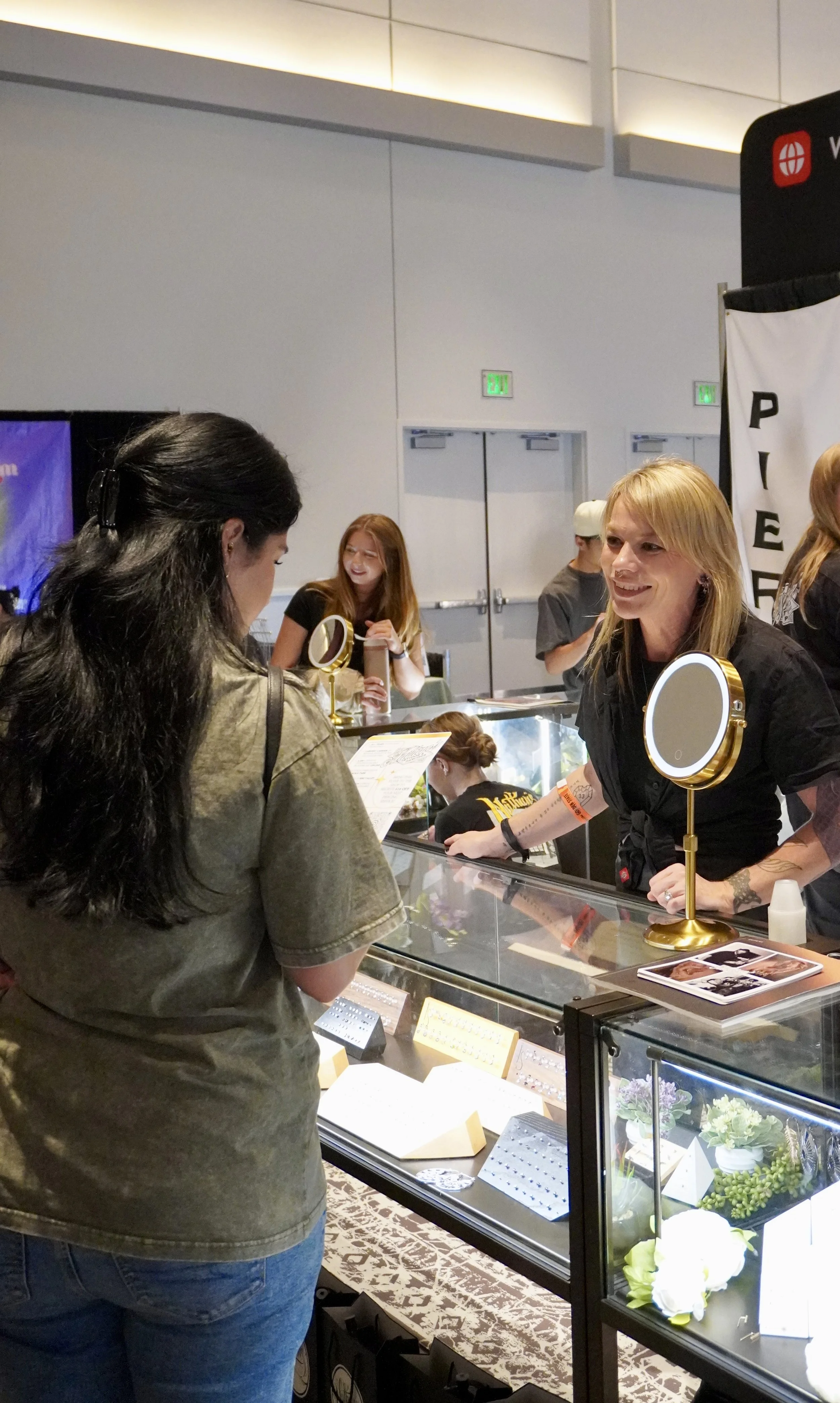 A woman shopping for jewelry at a display case with a jewelery vendor across the counter. The jewelry display contains rings, earrings, and decorative items, with a mirror on a stand and some small notes.