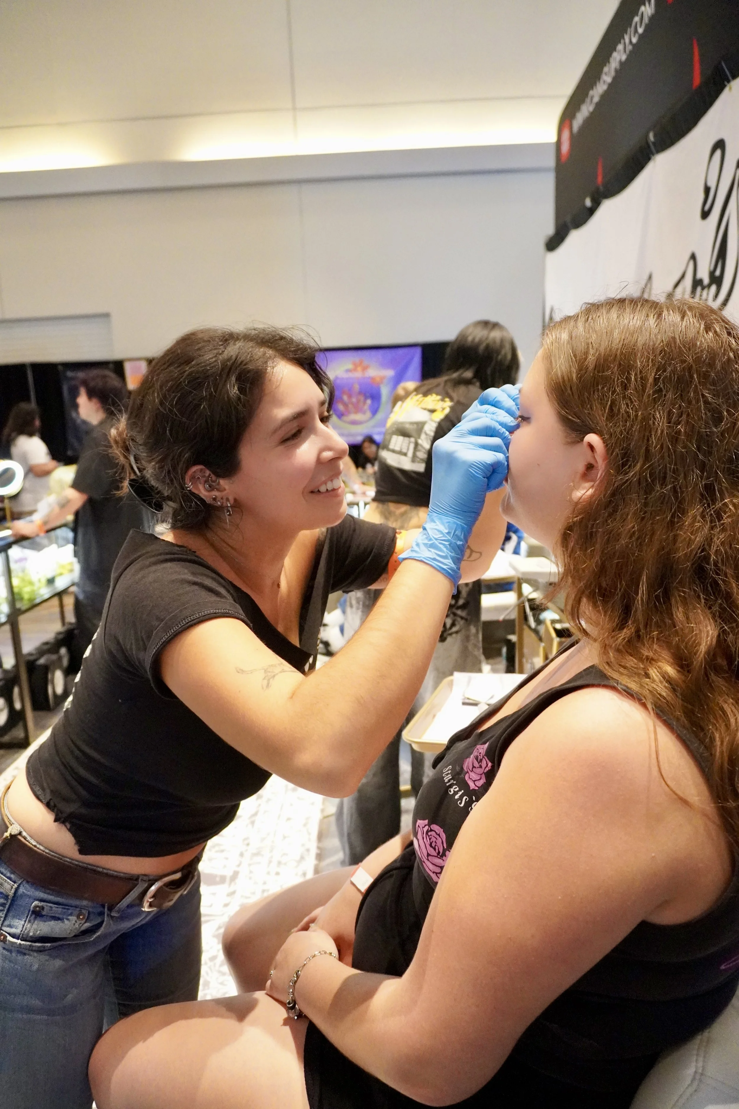 A woman with short brown hair, wearing a black shirt, is applying makeup near the face of a woman with curly red hair, who is seated, wearing a black tank top with pink floral designs. The woman with brown hair is smiling and wearing blue gloves. The