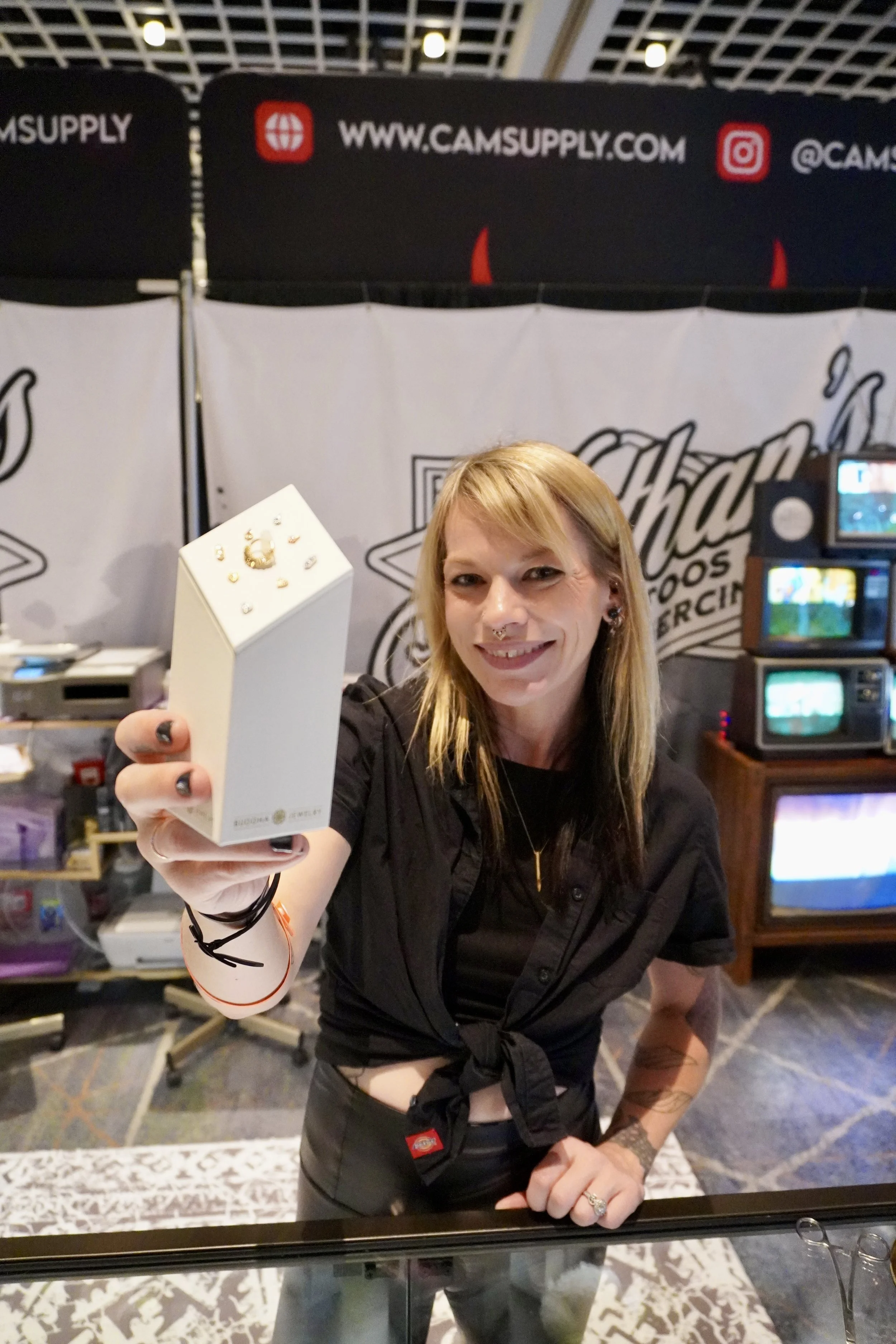 A smiling woman with blonde hair and tattoos on her left arm stands behind a glass counter, holding up a small box with jewelry inside. Behind her, there are banners with logos and website information, and shelves with electronic items and monitors i