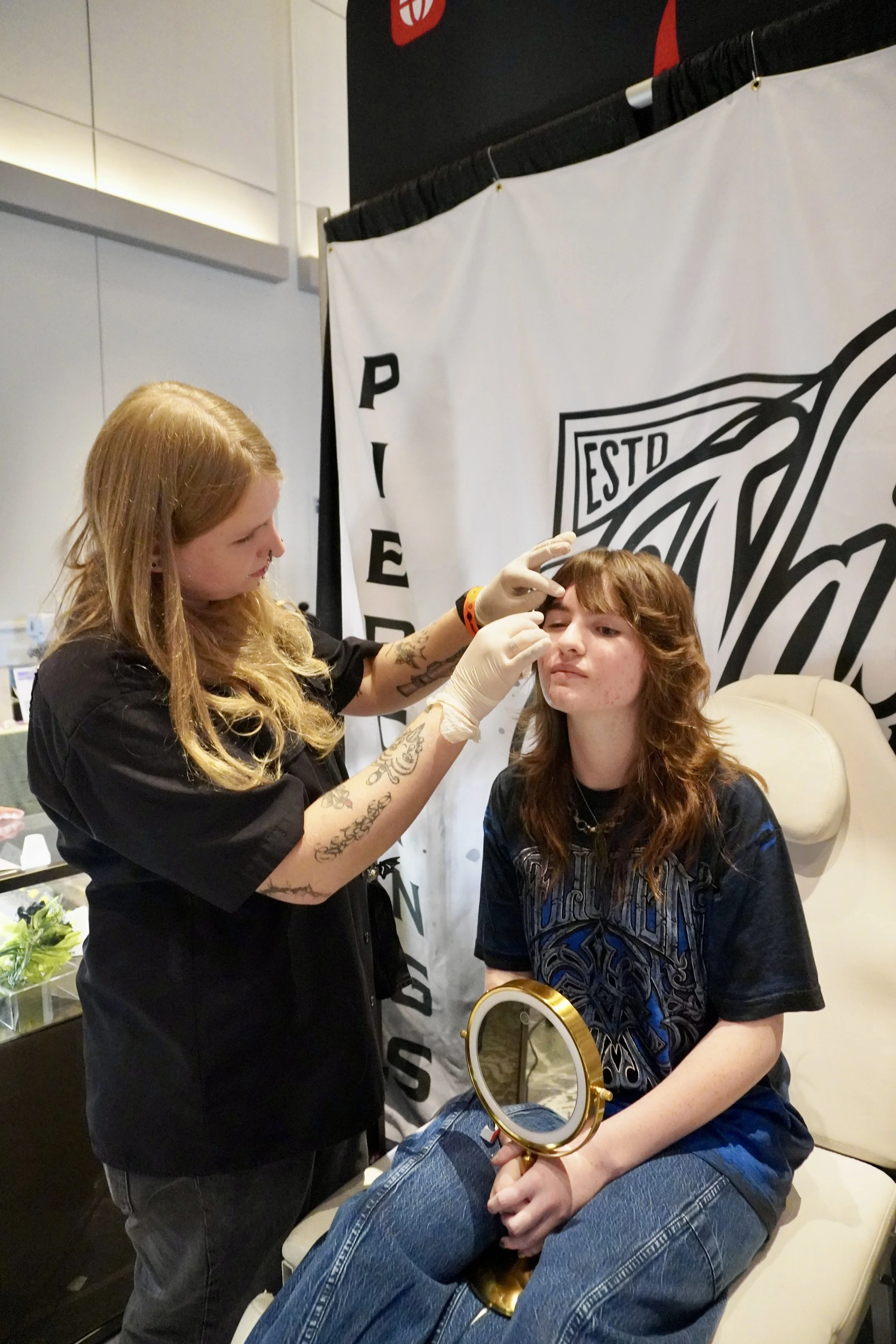 A woman with long reddish-brown hair is sitting in a chair while another woman with blonde hair, wearing gloves, appears to be giving her an eye cosmetic procedure. The woman in the chair is holding a round mirror, and both are indoors, with a banner