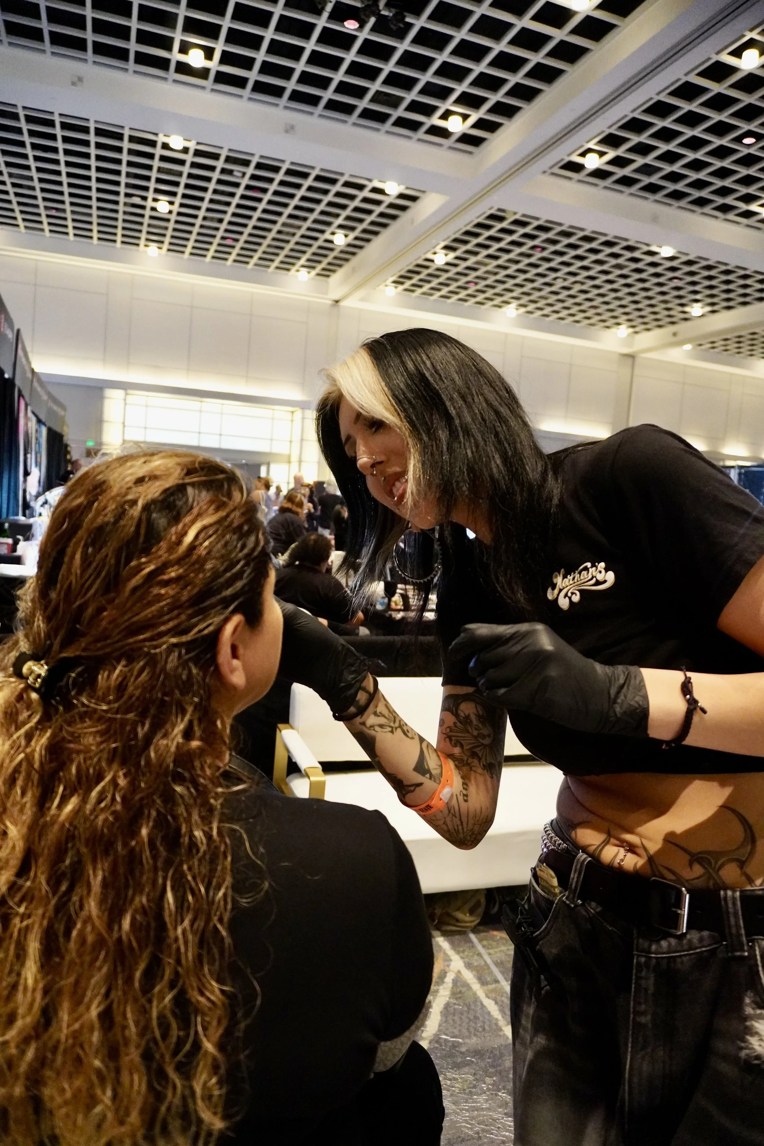 A tattoo artist with long black hair and tattoos on her arm, wearing a black crop top and black gloves, is tattooing a person's arm at a convention or event, with people in the background inside a large modern building with a patterned ceiling.