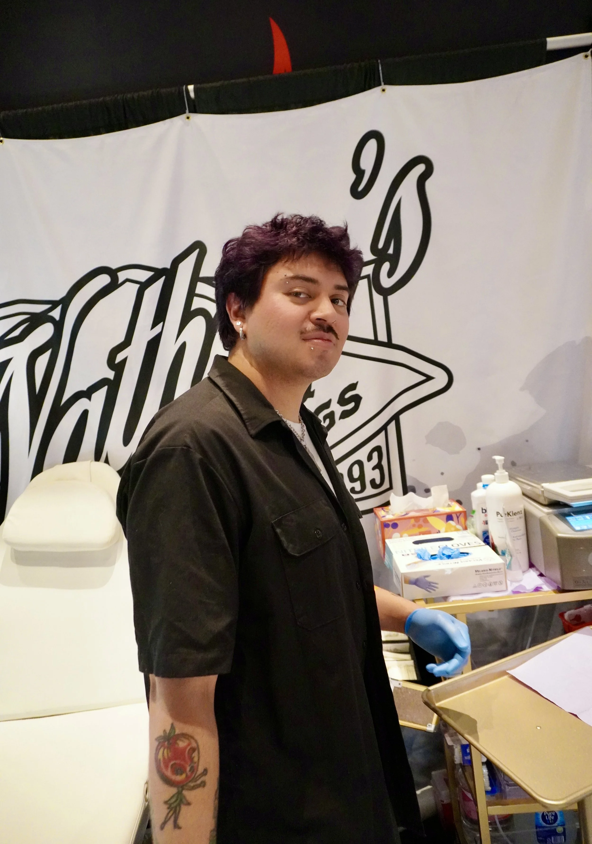 A young person with dark wavy hair and a facial piercing, wearing a black shirt, standing in a room with medical supplies on a table. There is a white wall with large black and white text and a graphic in the background.