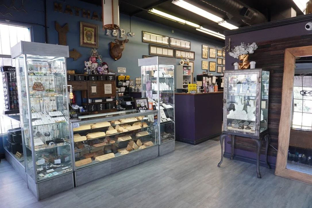 Interior of a jewelry store with glass display cases filled with jewelry, a purple cash register counter, framed artwork and decorations on the wall, and various display stands.